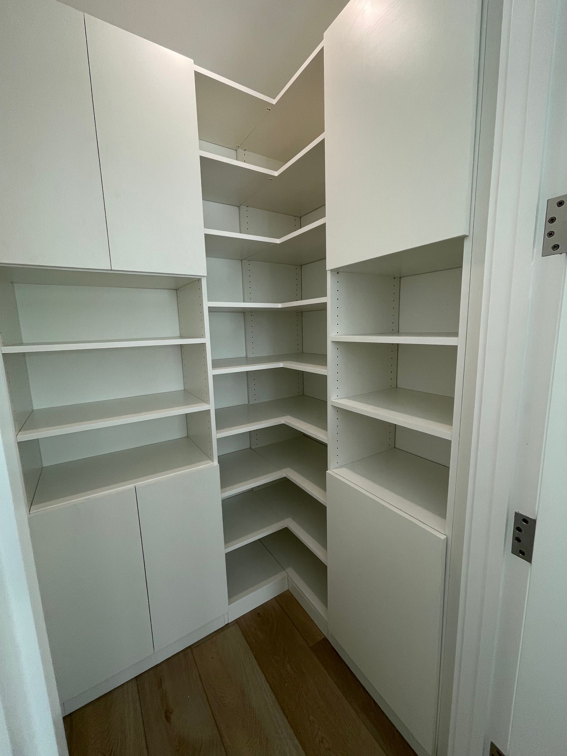 A corner walk-in pantry with white adjustable shelving units against light wood-look flooring.