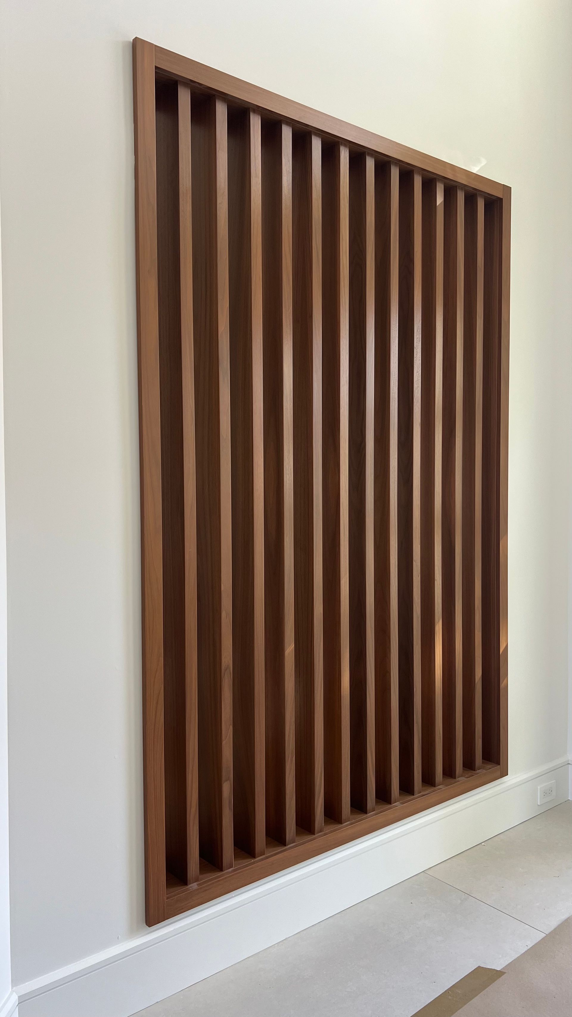 A rectangular wall-mounted wood slatted screen with vertical panels framed in a matching wood finish against a white wall.