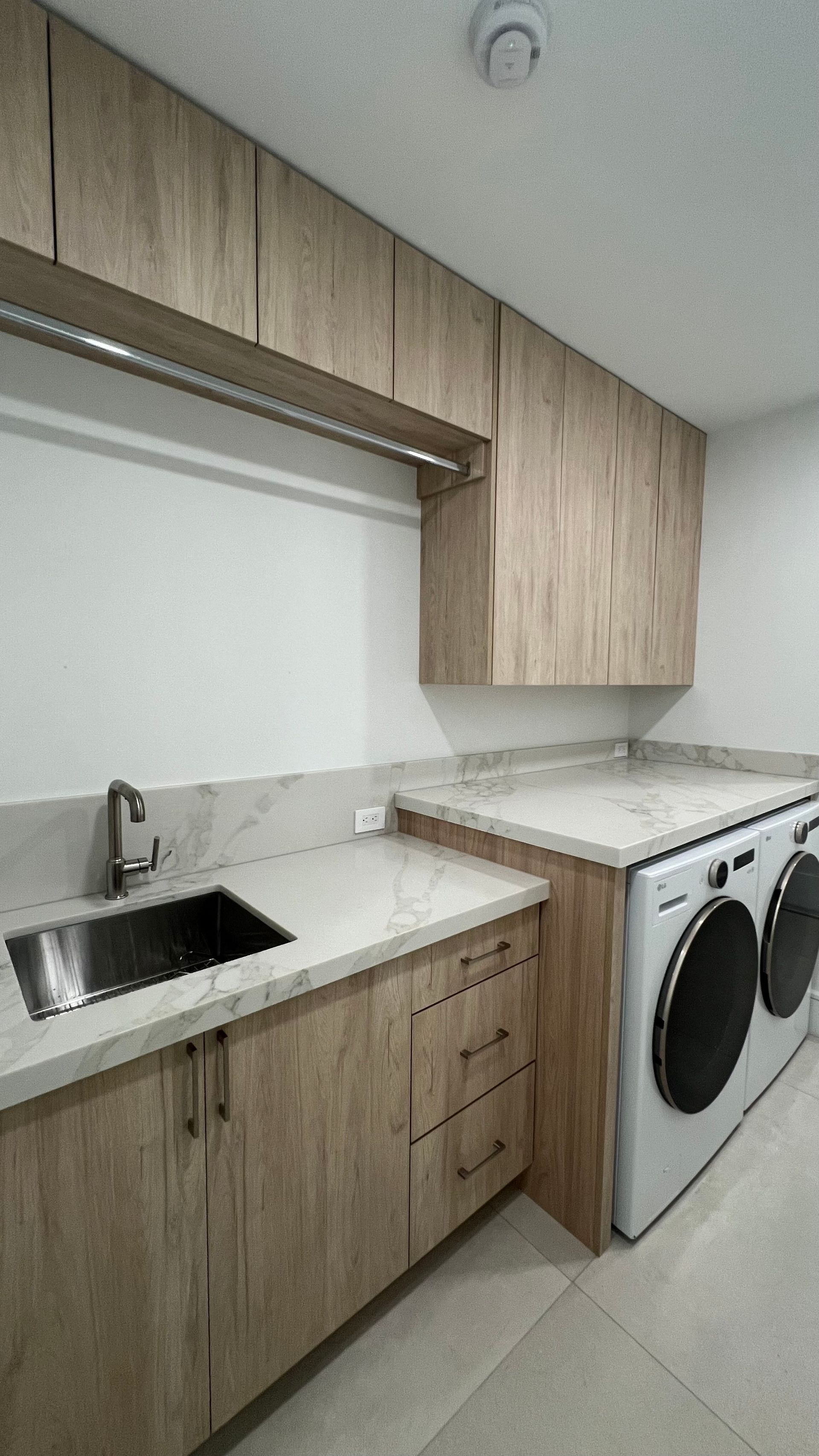 A modern laundry room with wood-grain cabinets, a stainless steel sink, light countertops, and a white washing machine.