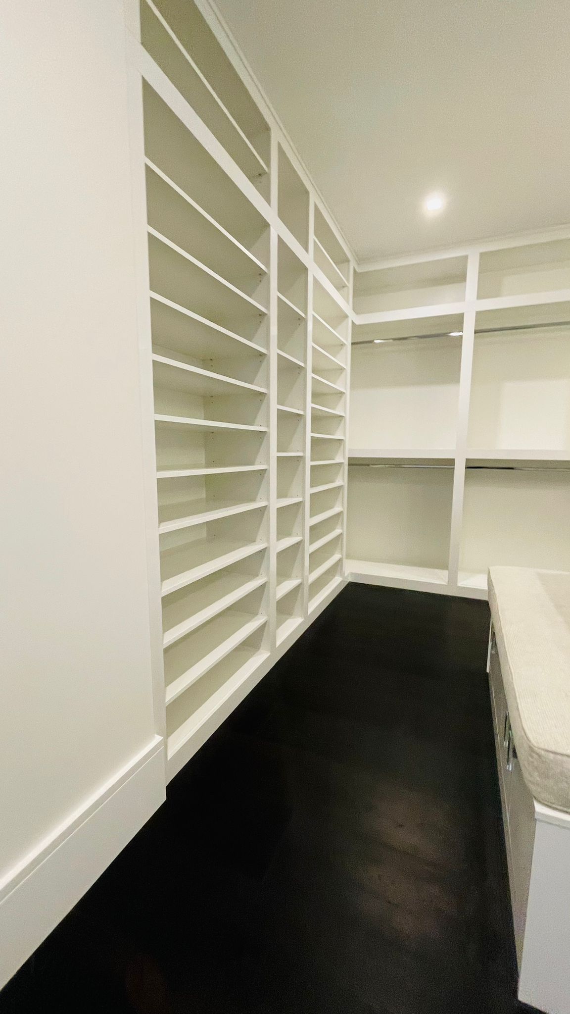 A spacious walk-in closet featuring floor-to-ceiling white shelving, an island counter, and dark flooring.