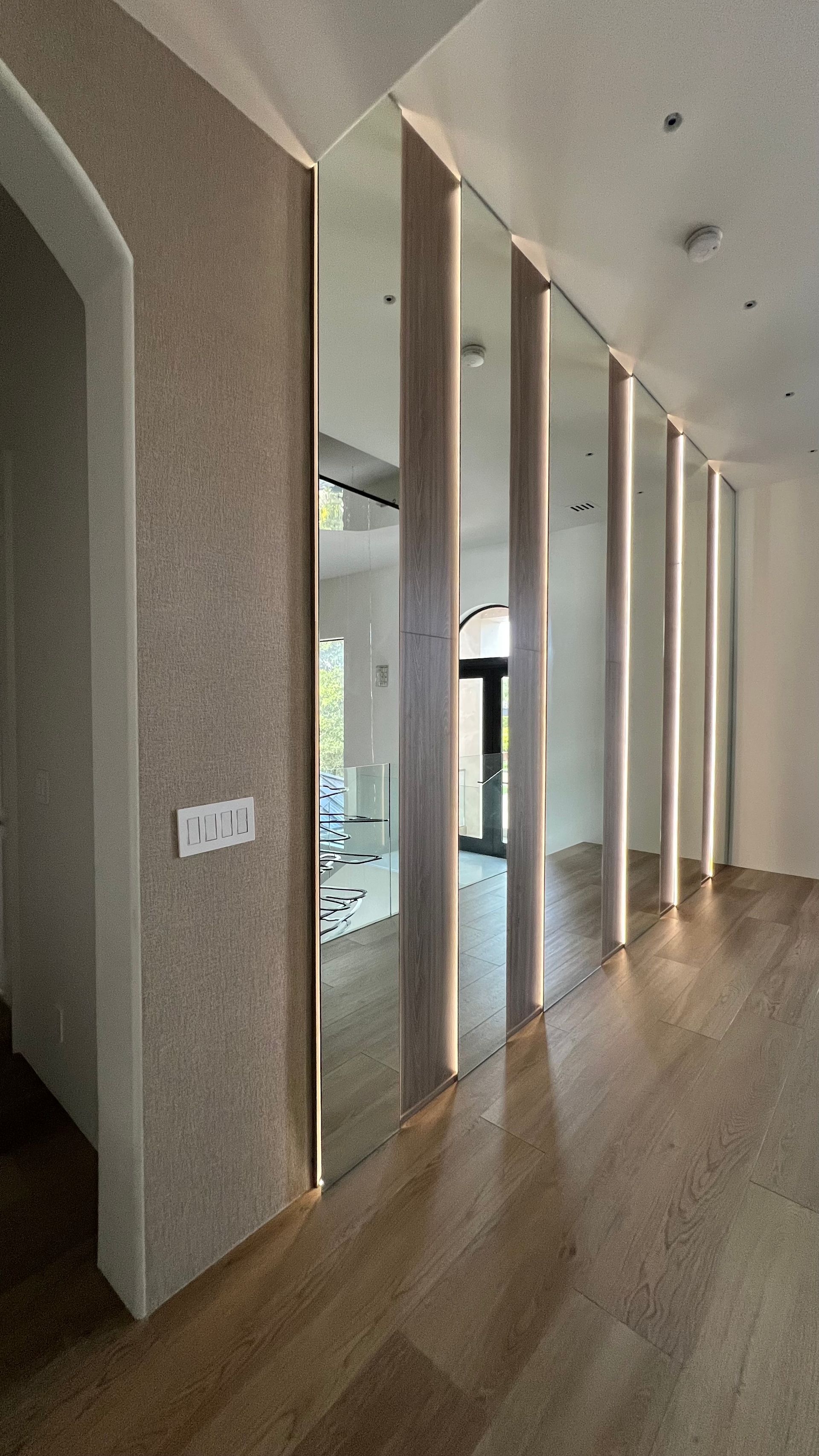 A hallway wall with vertical mirror panels separated by illuminated wood strips above light wood flooring.