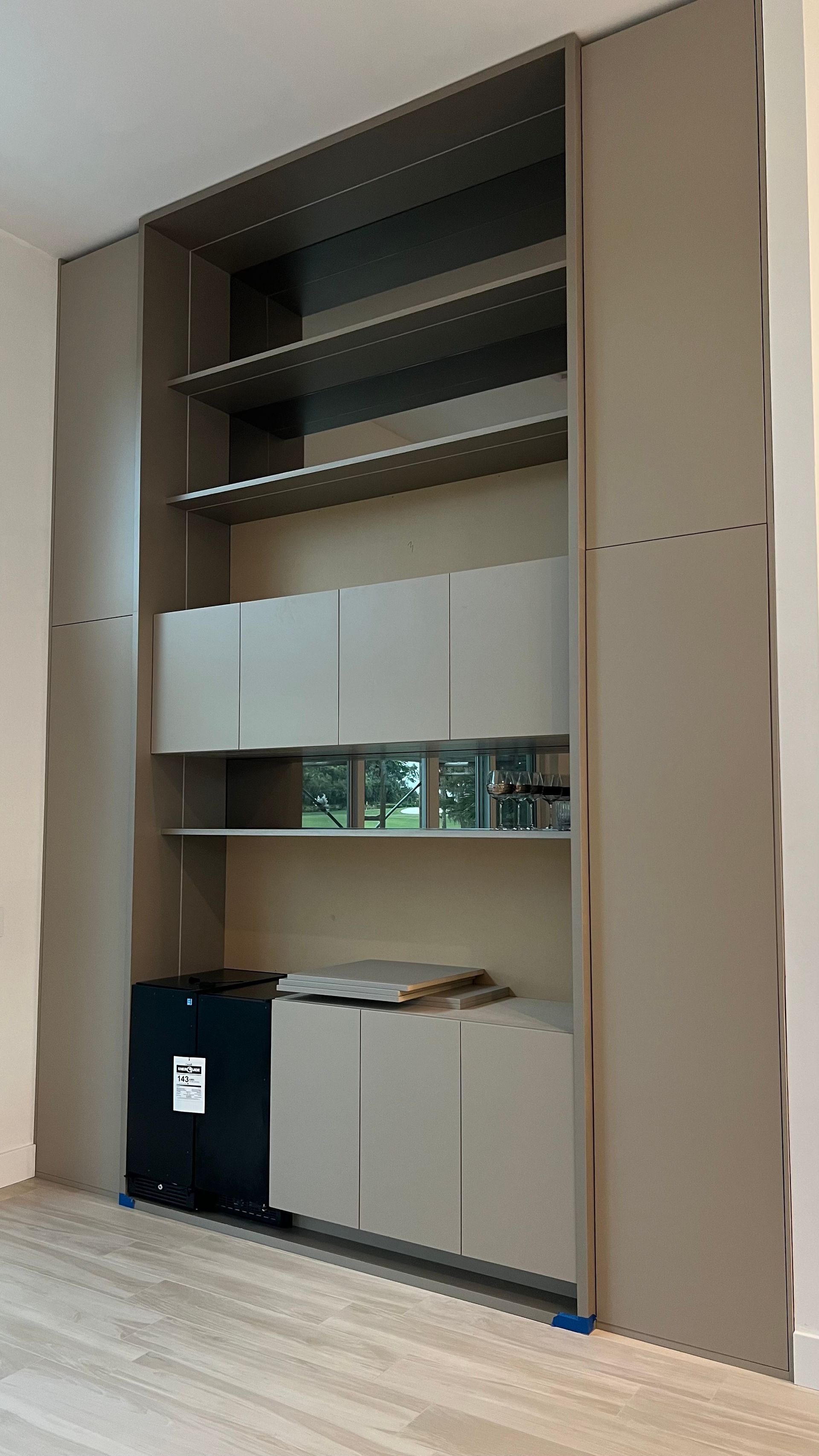 A modern floor-to-ceiling built-in wall unit with shelving, light gray cabinets, and a dark under-counter refrigerator.