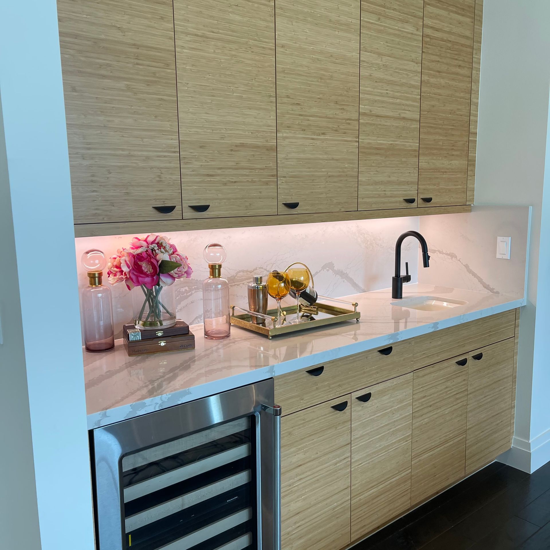 A modern wet bar featuring light wood cabinets, white marble countertops, a wine fridge, and decorative glass accents.