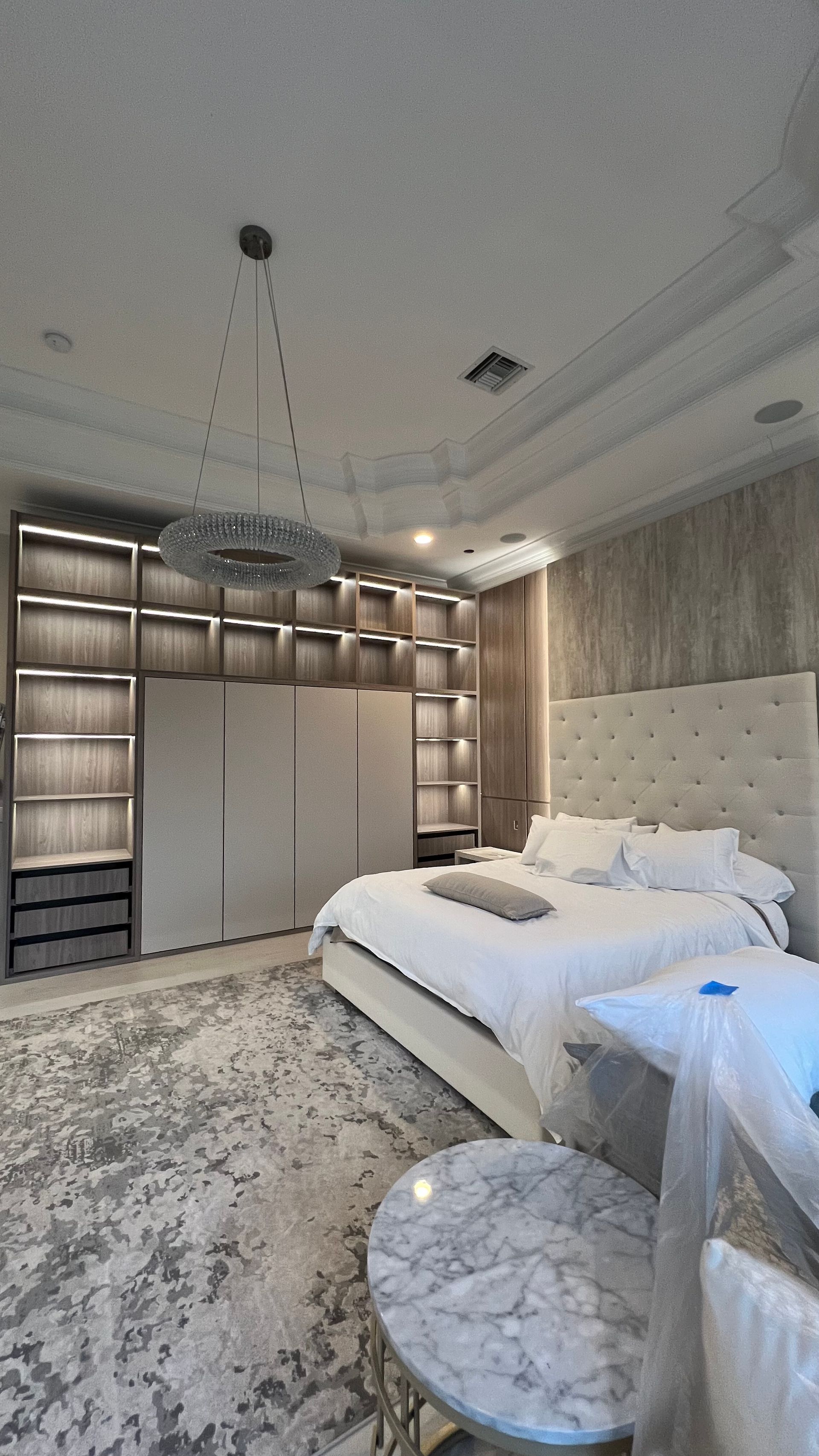 A modern, luxurious bedroom with a large bed, plush white bedding, wall-to-wall cabinetry, and a circular marble table.