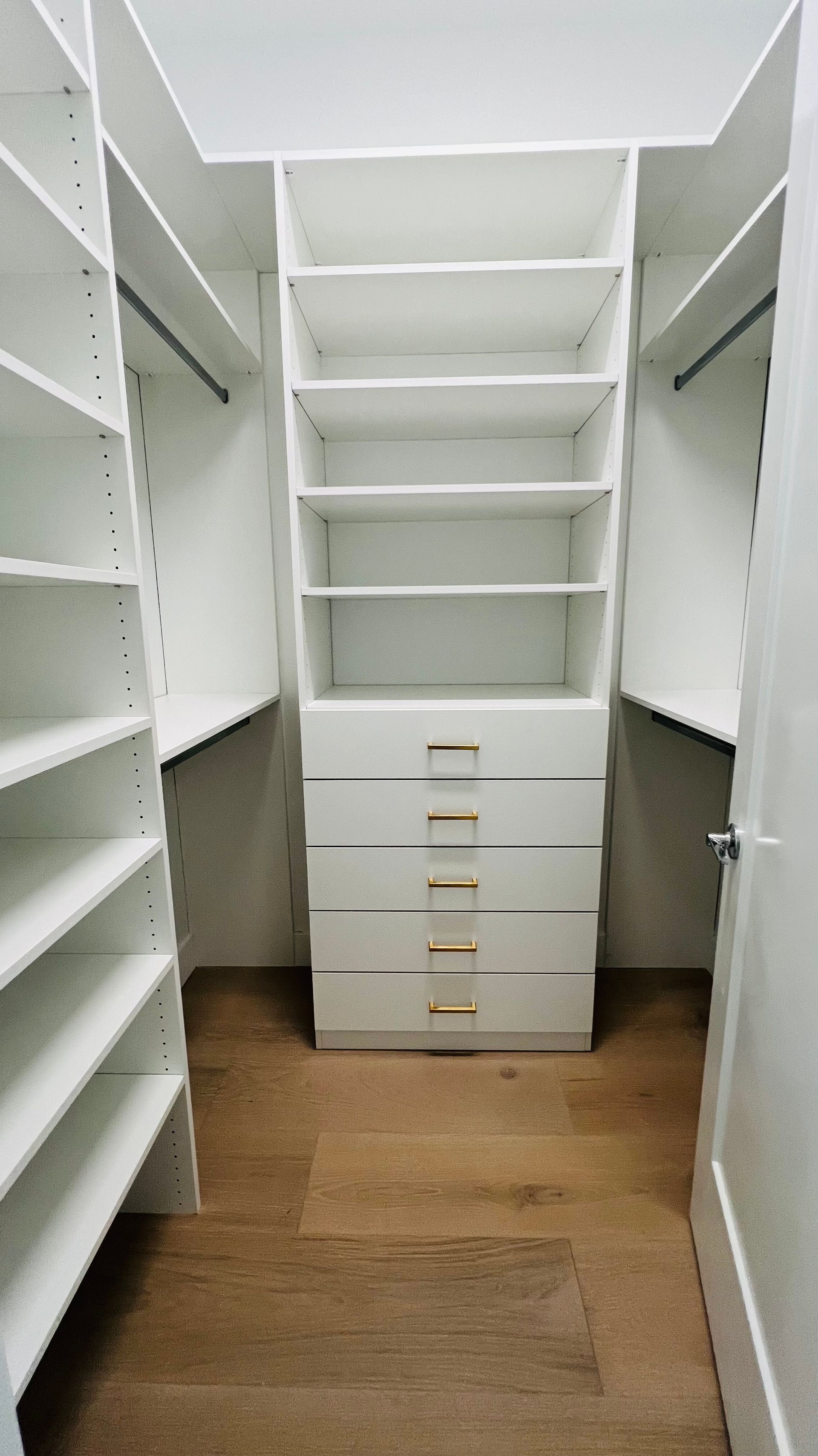 A walk-in closet featuring white shelves, hanging rods, and a central chest of five drawers on a hardwood floor.