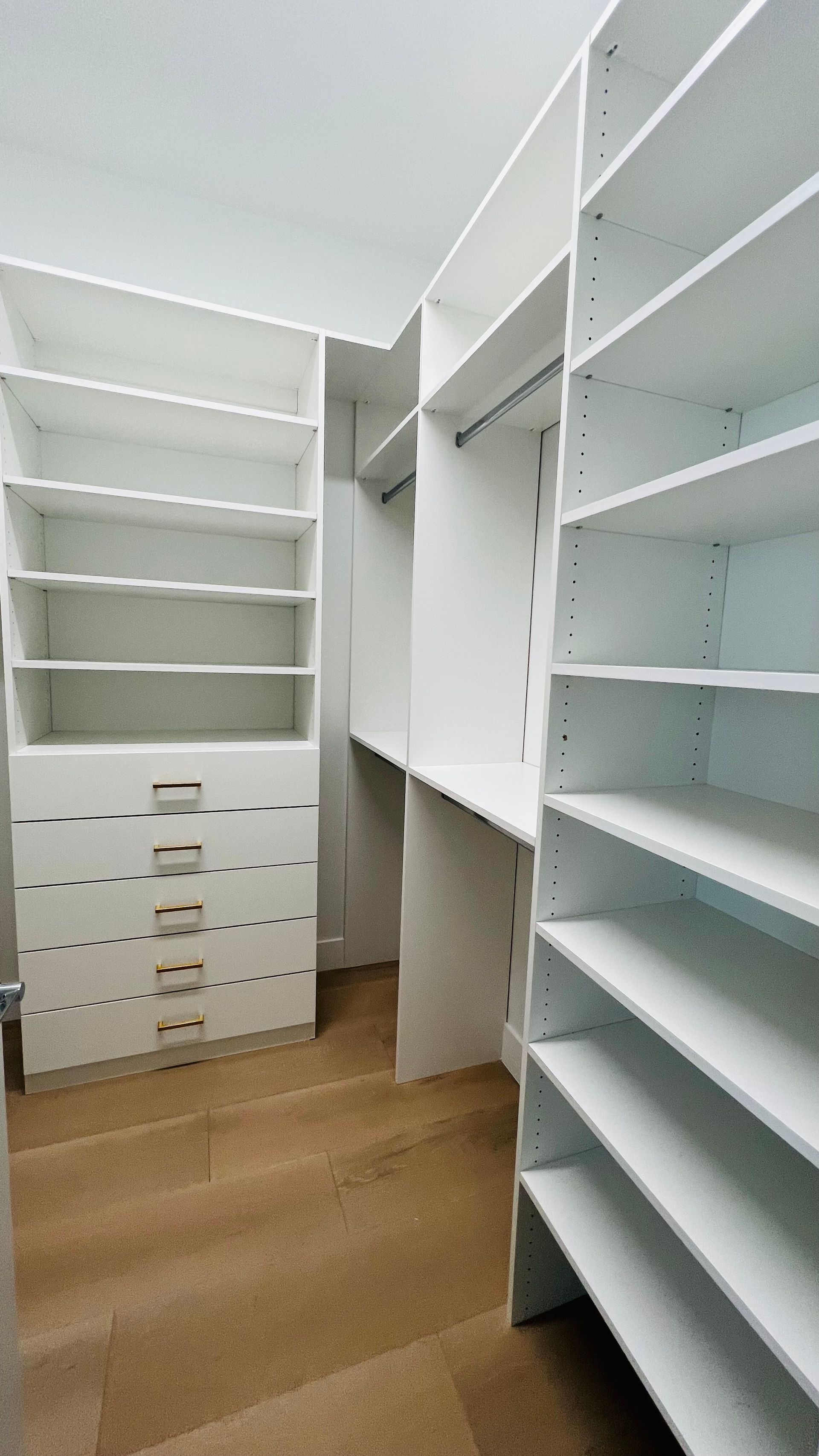A walk-in closet with white shelving, a stack of five drawers, and hanging rods against wood-look flooring.