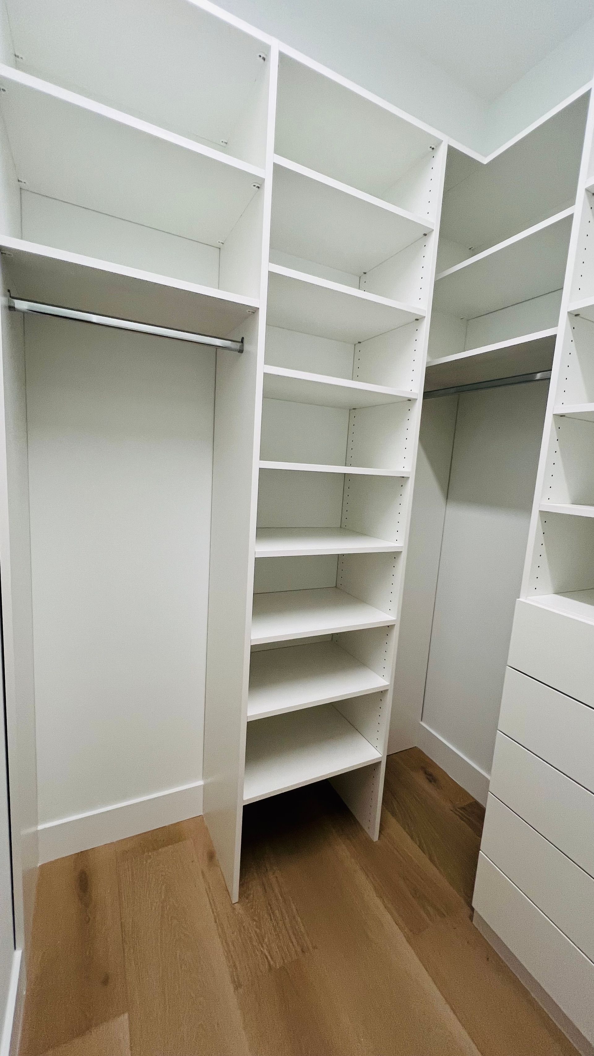 A walk-in closet featuring white wooden shelving, a hanging rod, and drawers, set against a light wood floor.