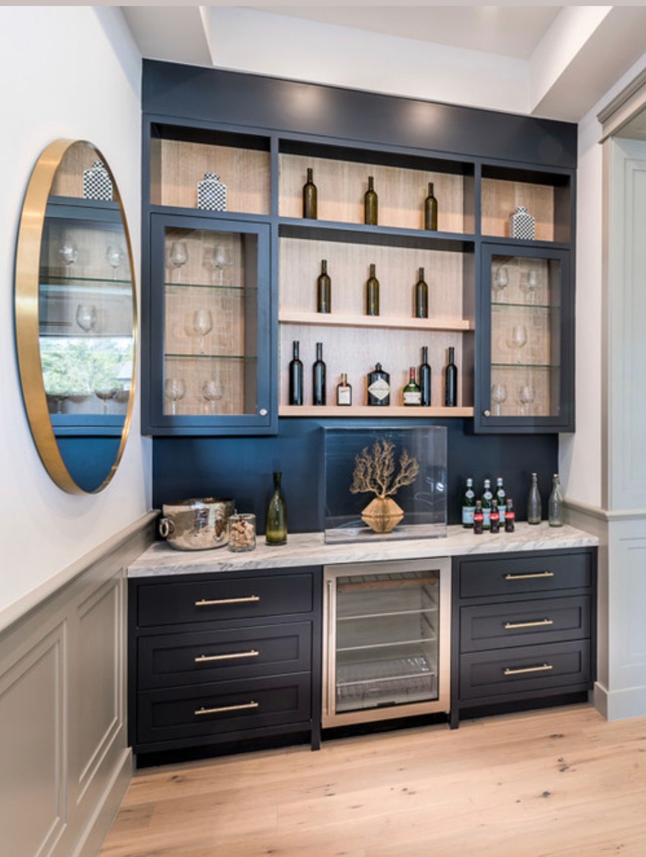 A dark blue custom home bar with light wood shelves, glassware, bottles, a marble countertop, and a wine cooler.
