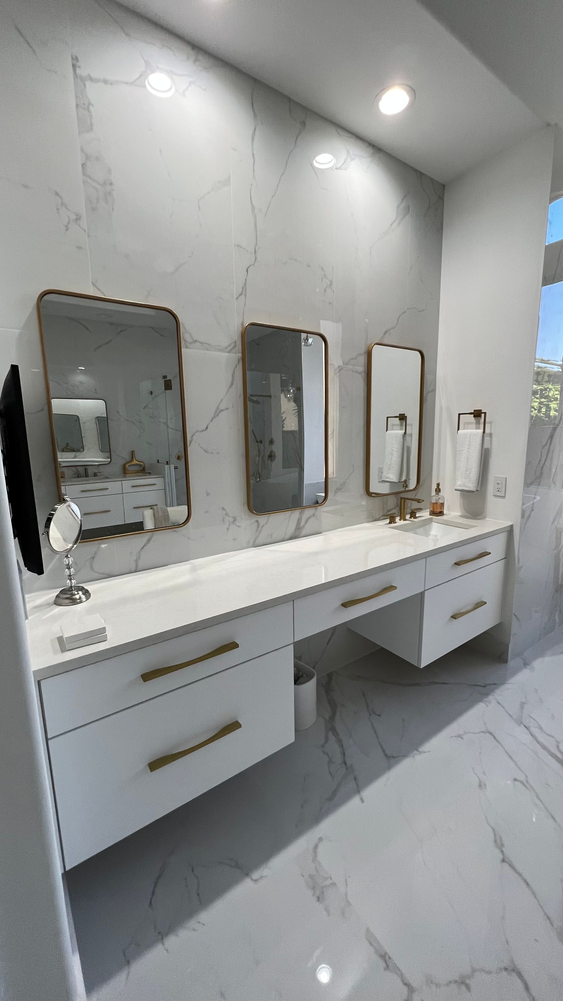 Modern floating bathroom vanity with white marble walls, two gold-trimmed mirrors, and gold hardware on white cabinets.