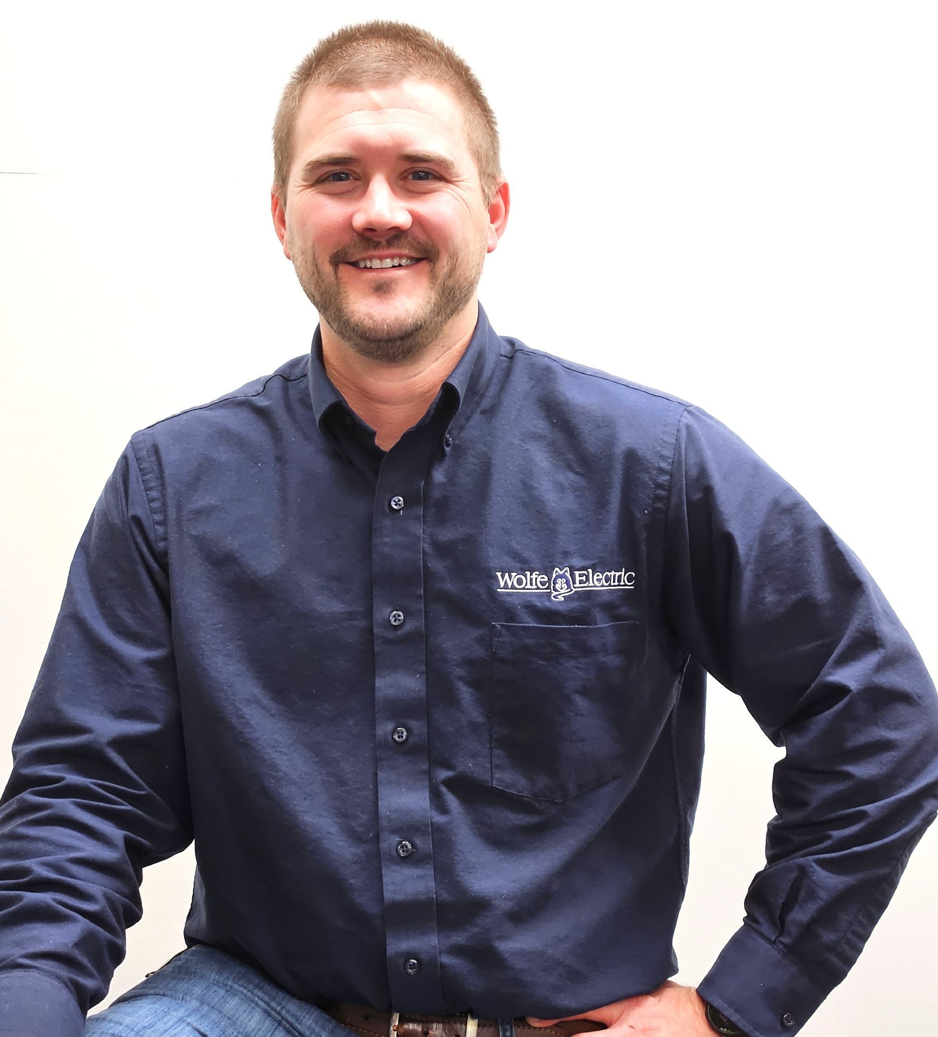 Man with a beard in a blue button-down shirt smiling, hand on hip, against a white backdrop.