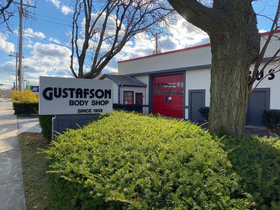 Gustafson Body Shop sign in front of a gray building with a red garage door, tree and bushes.