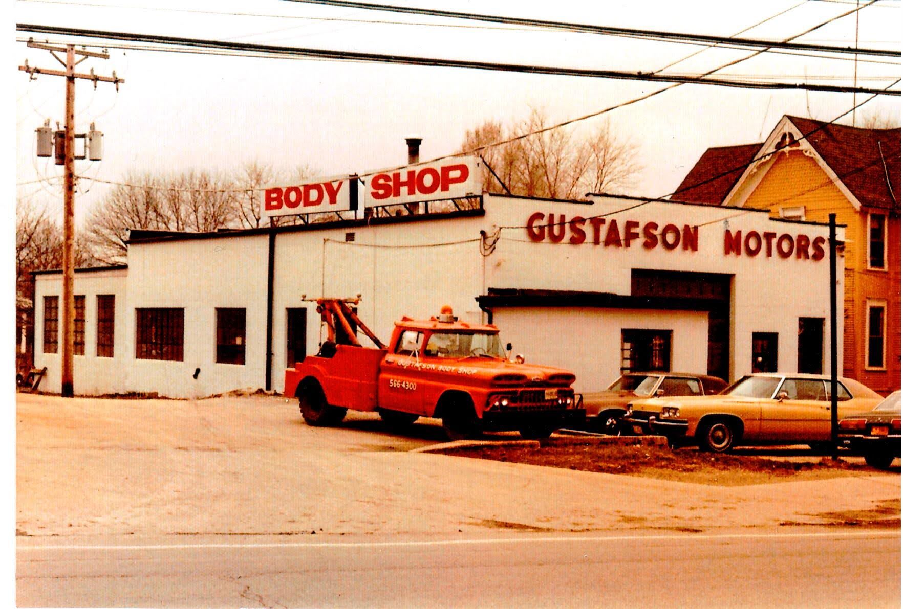 Red tow truck parked outside "Gustafson Motors" and "Body Shop" buildings. Several cars are parked nearby.
