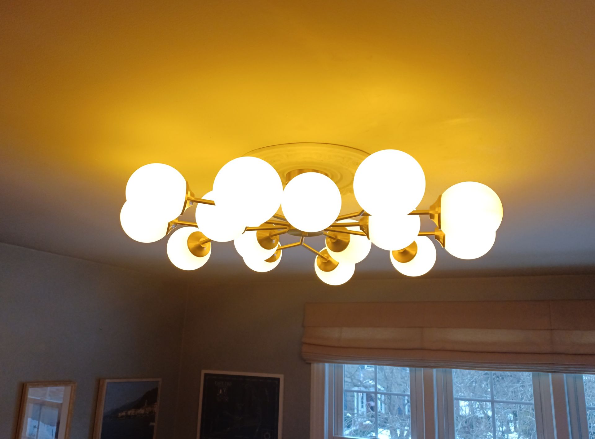 A golden chandelier with globe lights illuminates a room, with a window and artwork visible.