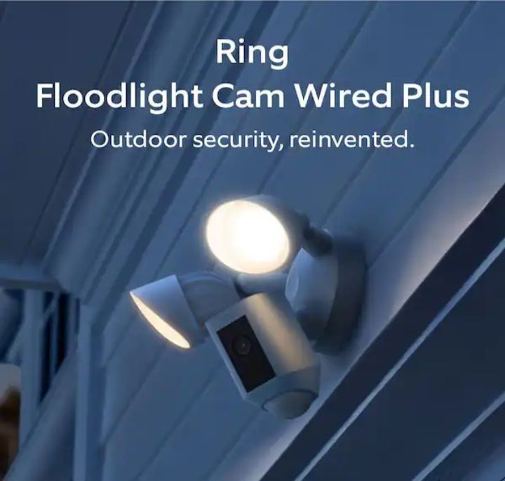 Ring Floodlight Cam Wired Plus mounted on blue siding, with bright floodlight on outdoor security camera device