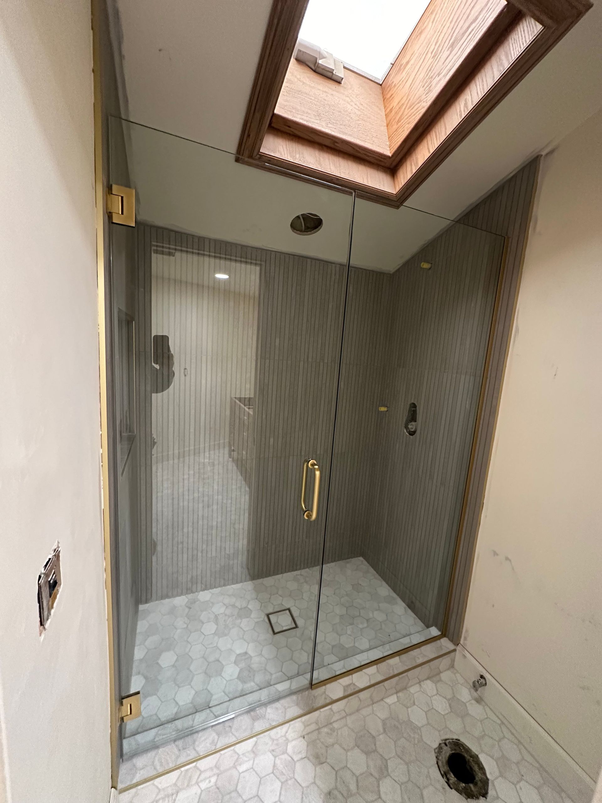 Shower stall with glass door, gold hardware, grey tile, and a skylight.