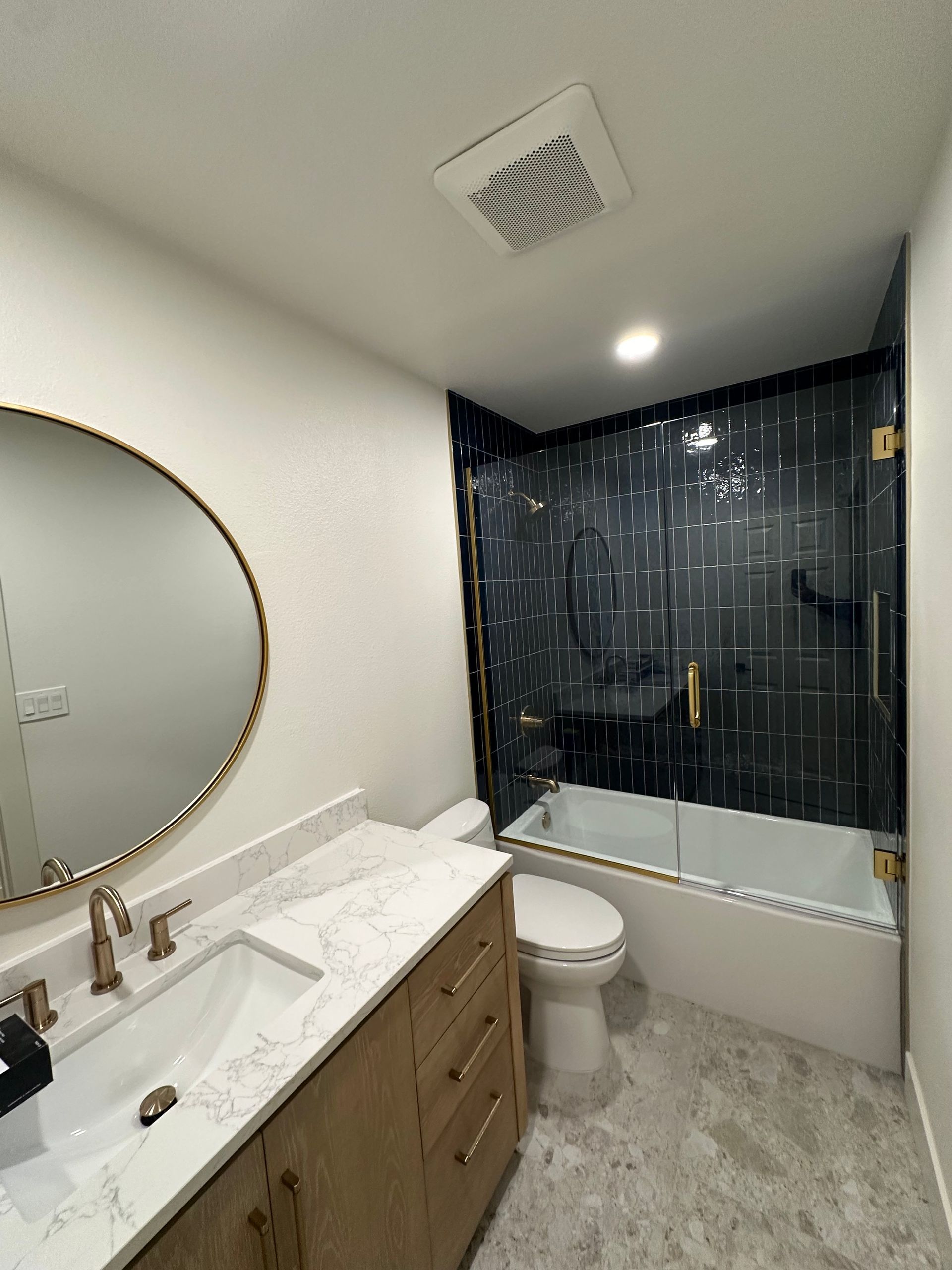 Shower with glass door, gold hardware, grey tile walls, and a skylight.