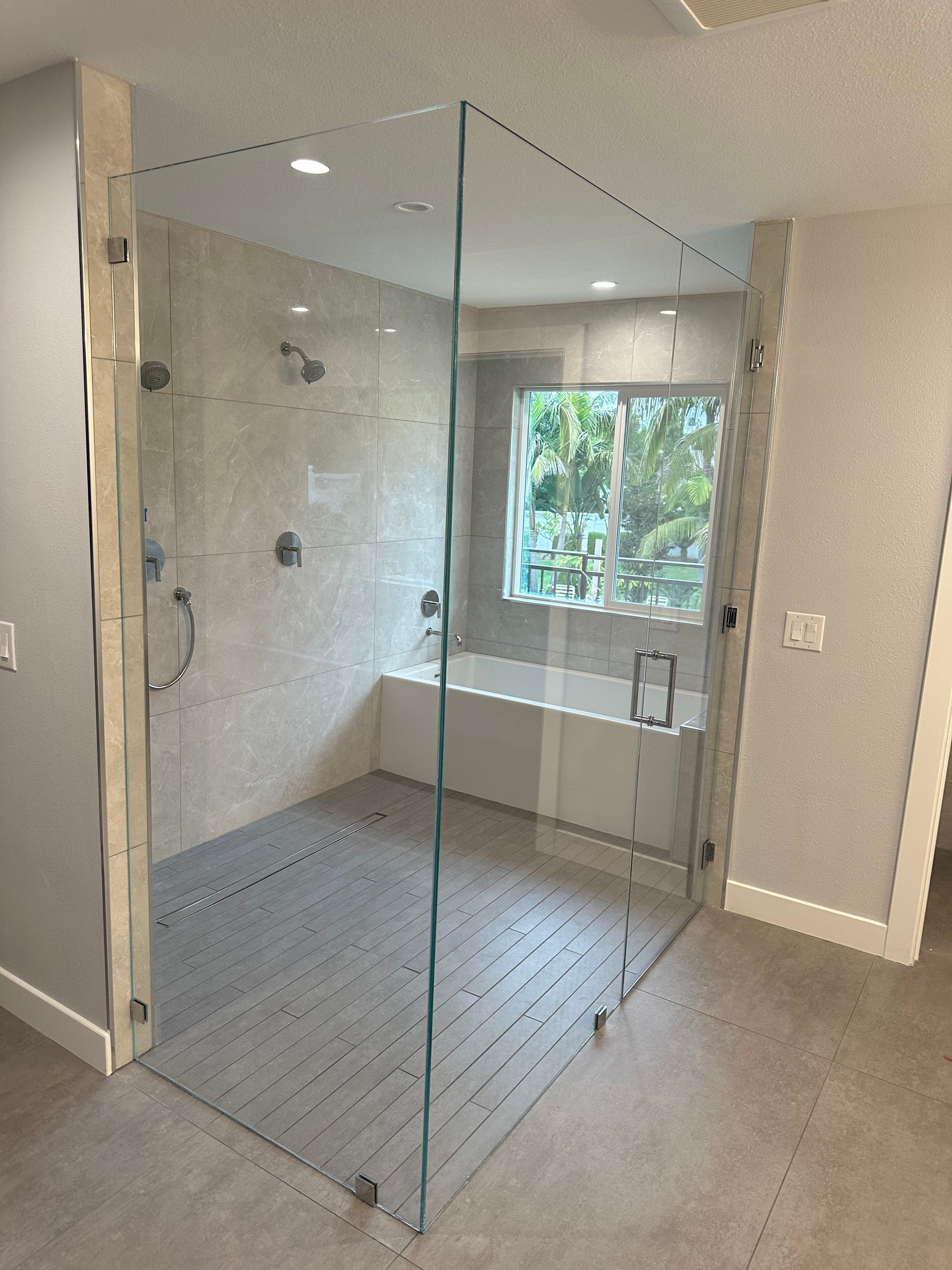 Glass-walled shower with white tiled walls, shower head, and a bathtub with a window view in the background.