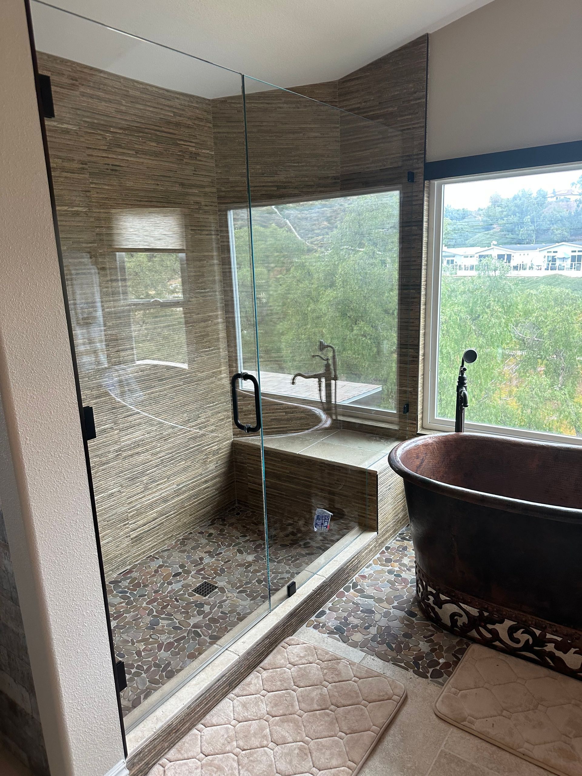 Shower with pebble tile walls, glass door, and copper tub by a window with a natural view.