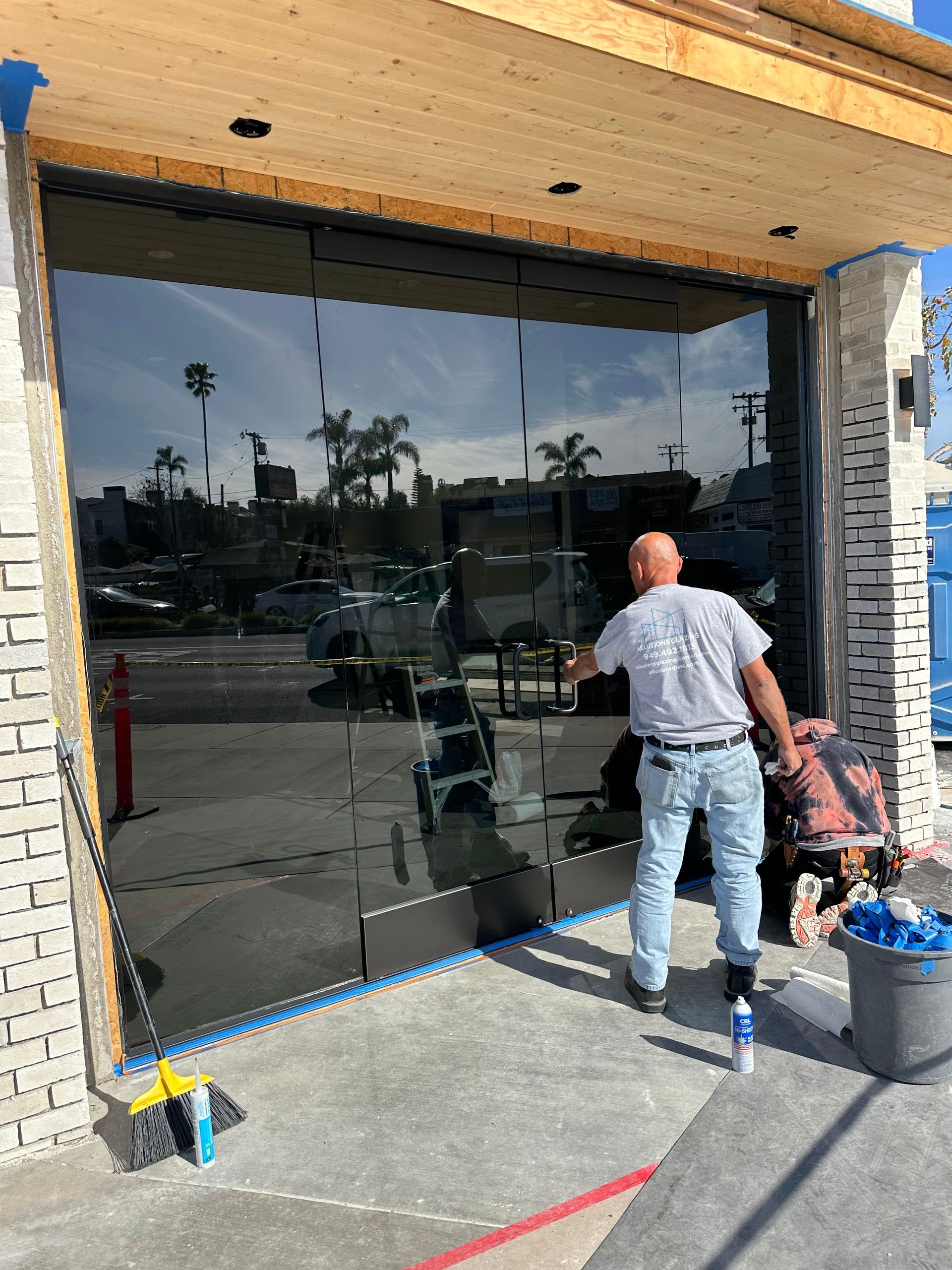 Man cleaning a large, tinted glass storefront. Sunlight reflects off the glass, a brush and bucket are nearby.