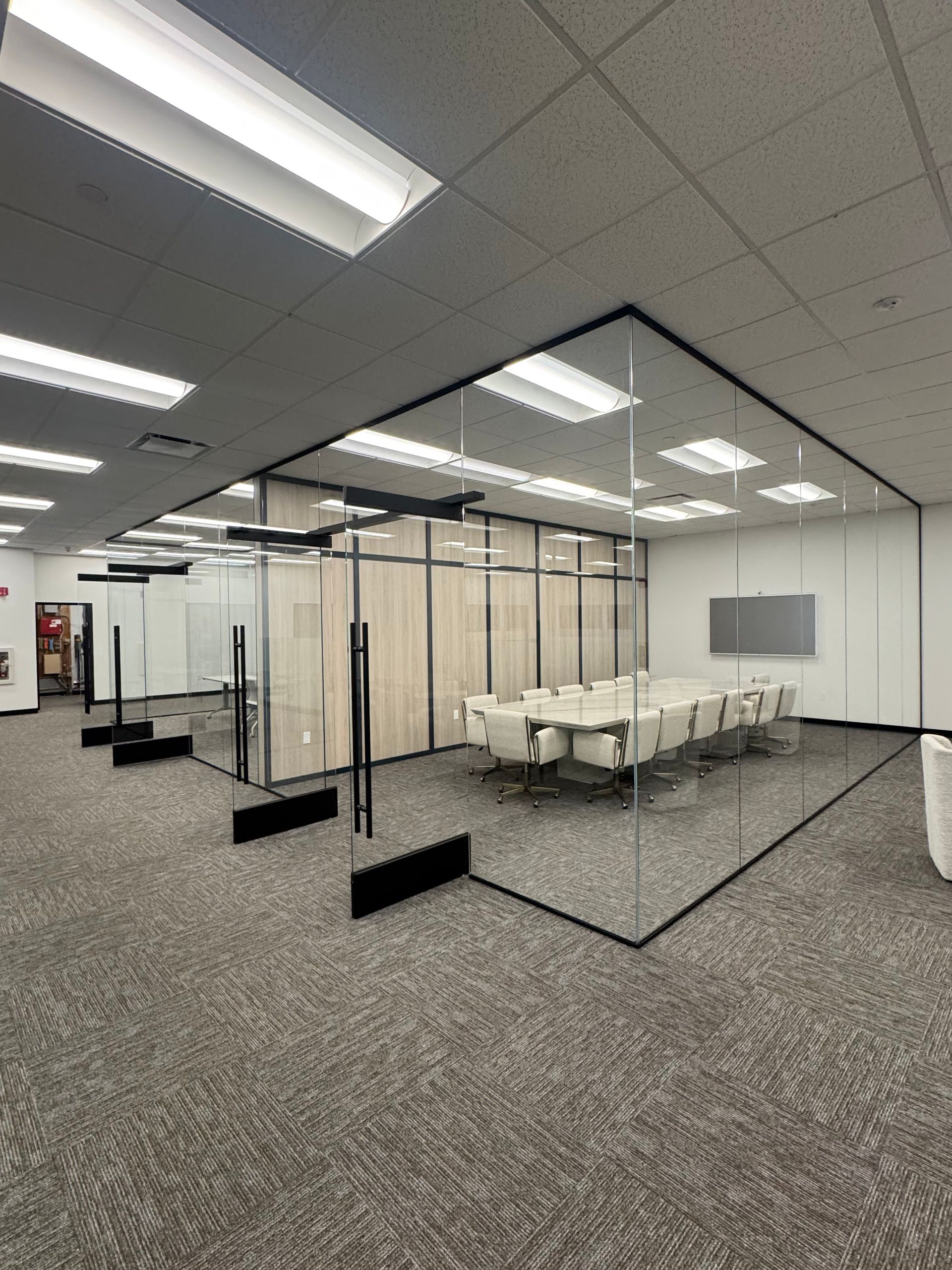 Conference room with glass walls, a long table, and a light-colored wood paneling, carpeted floor, and overhead lighting.
