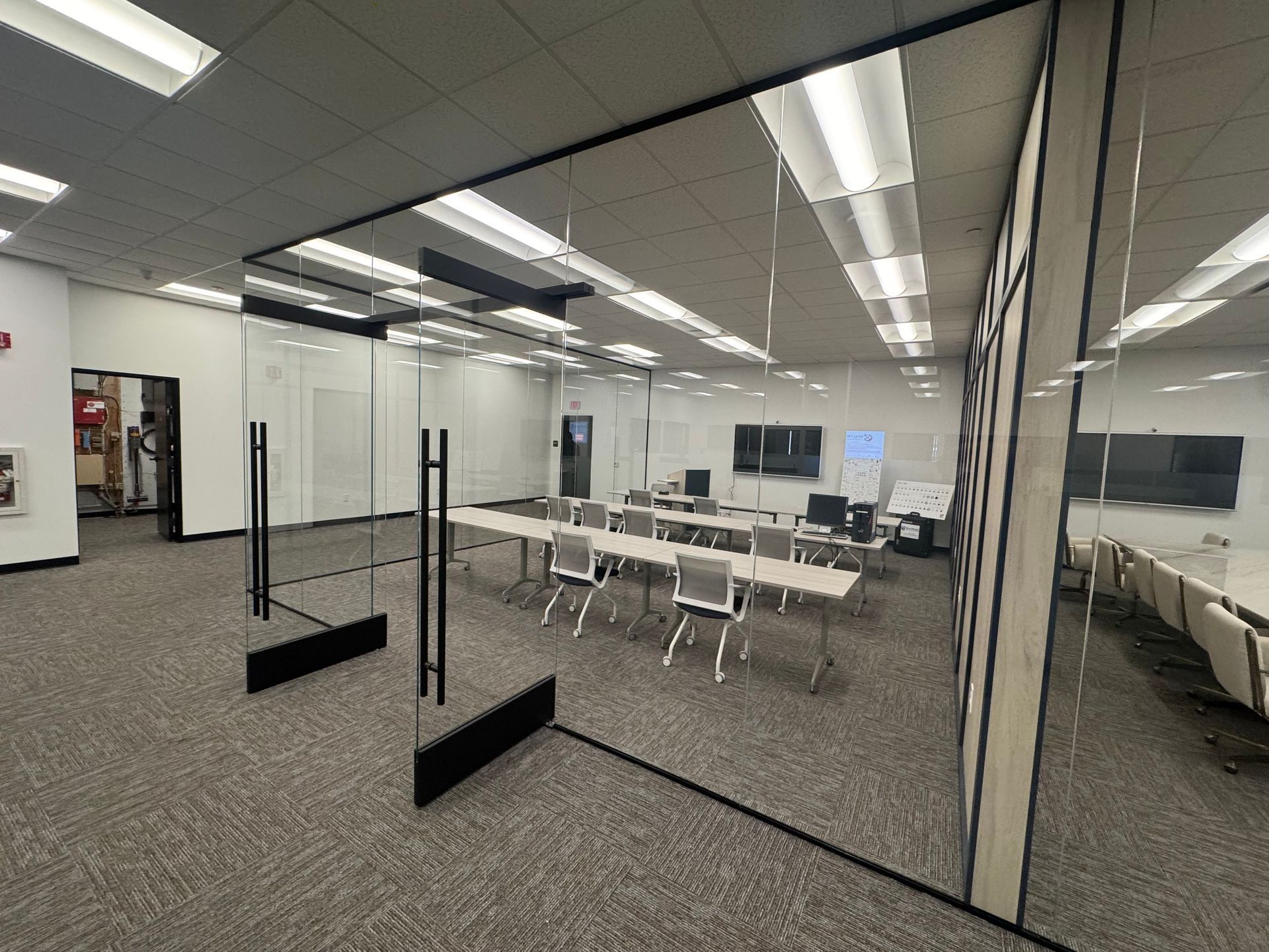 Conference room with glass walls and tables, chairs, and multiple monitors. Black dividers in foreground.