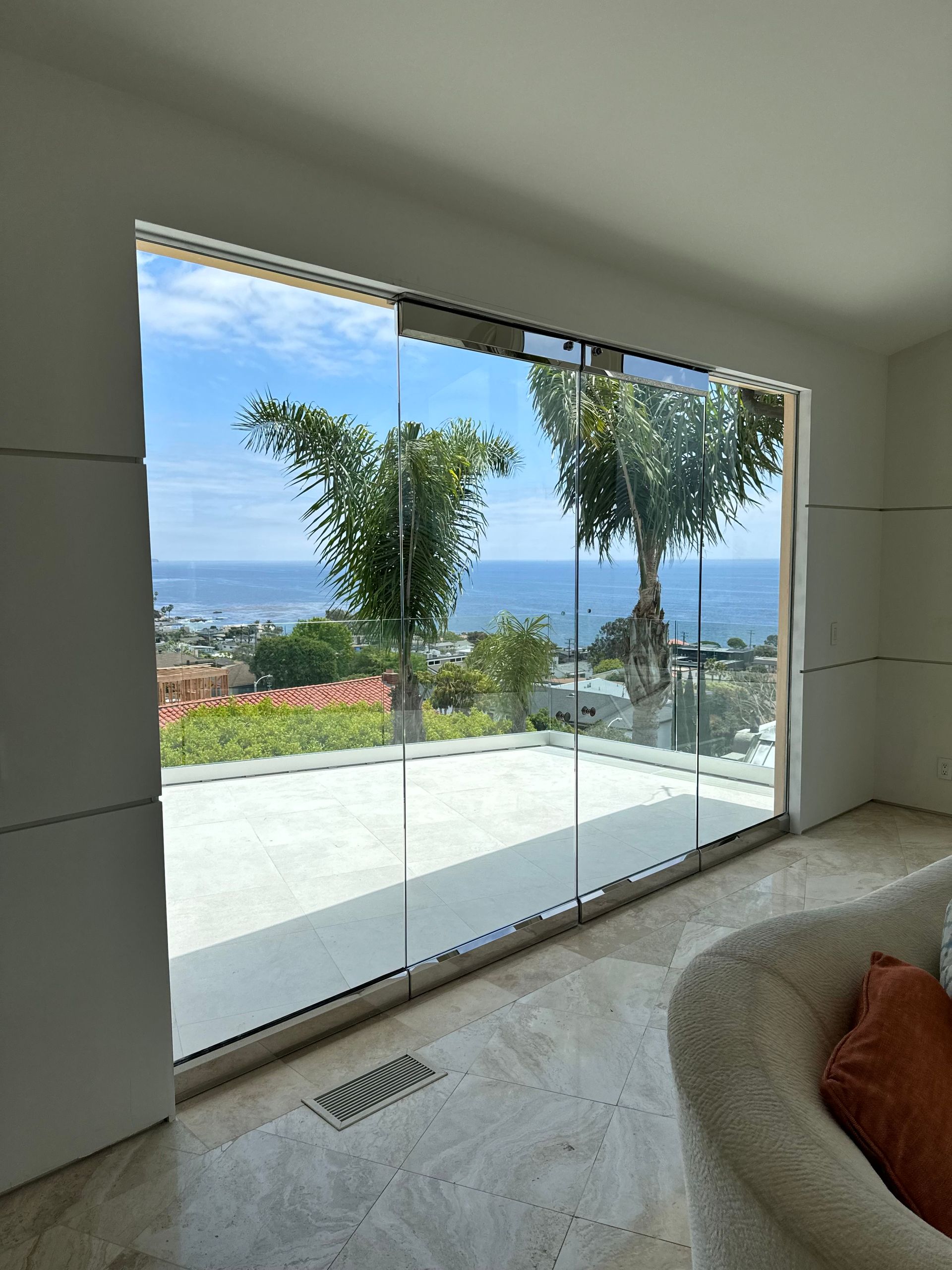 Large glass window with ocean view, trees, and sunny sky. Interior with white walls and a tan chair.