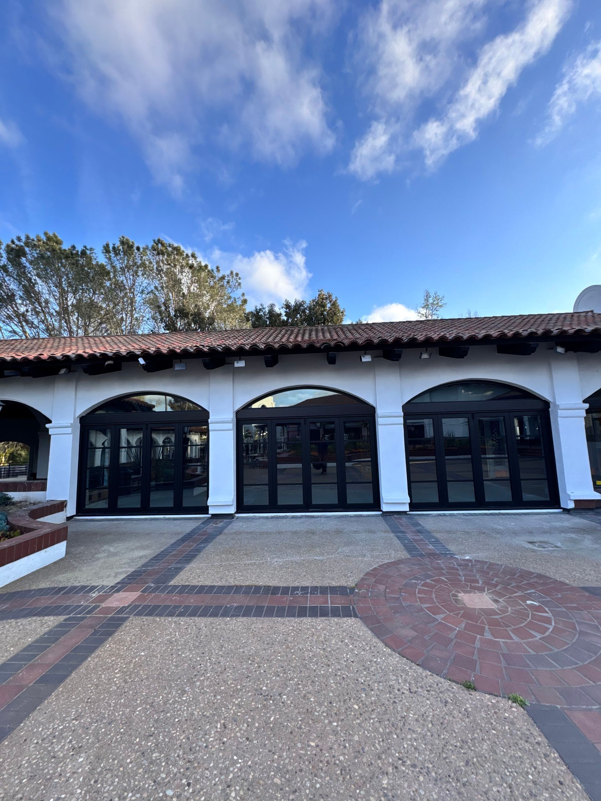 Exterior view of a white arched building with black folding doors, red tile roof, brick and concrete patio. Cloudy blue sky.