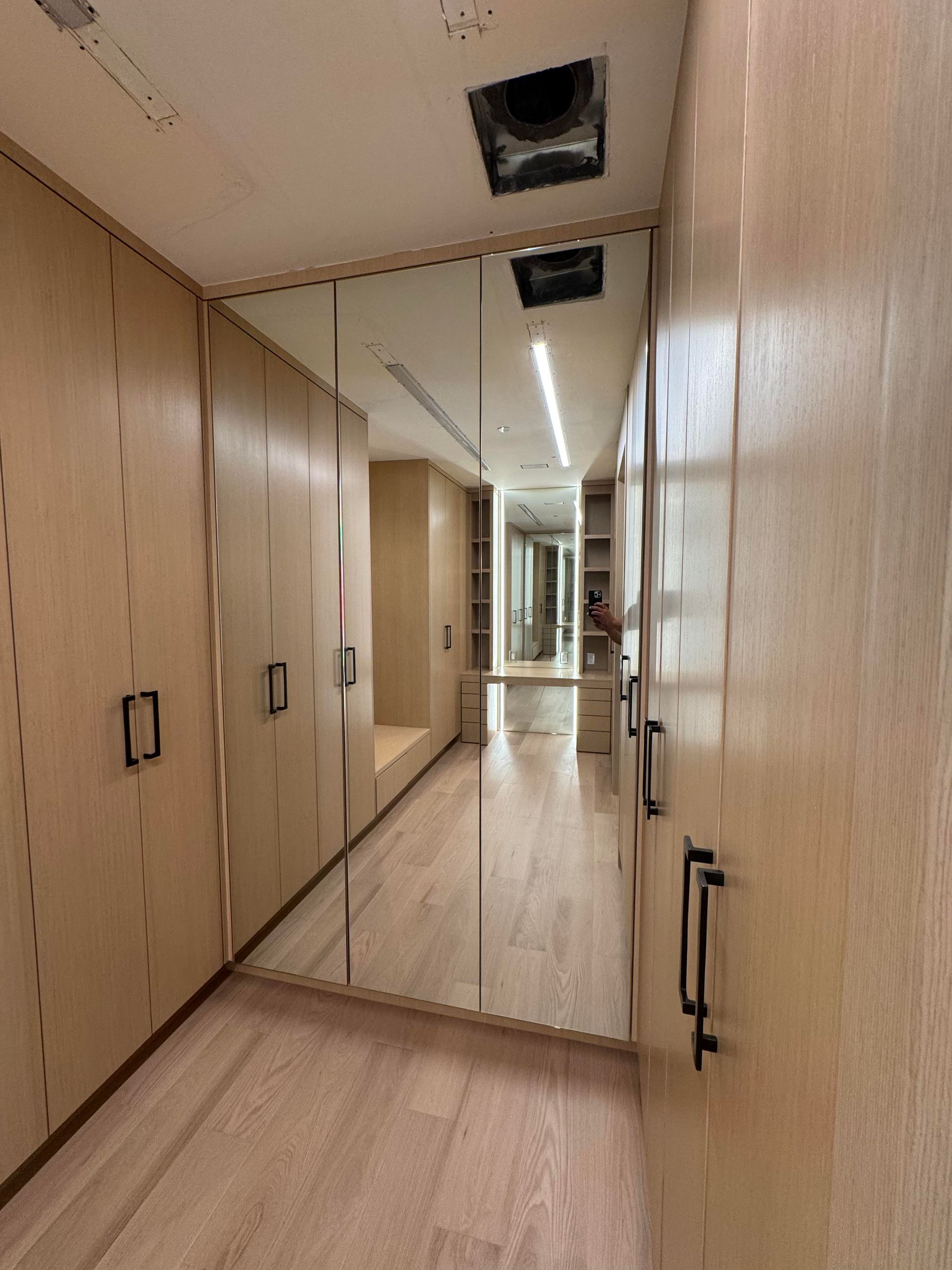Narrow walk-in closet with light wood cabinets, long mirror, light wood floors, and black hardware.