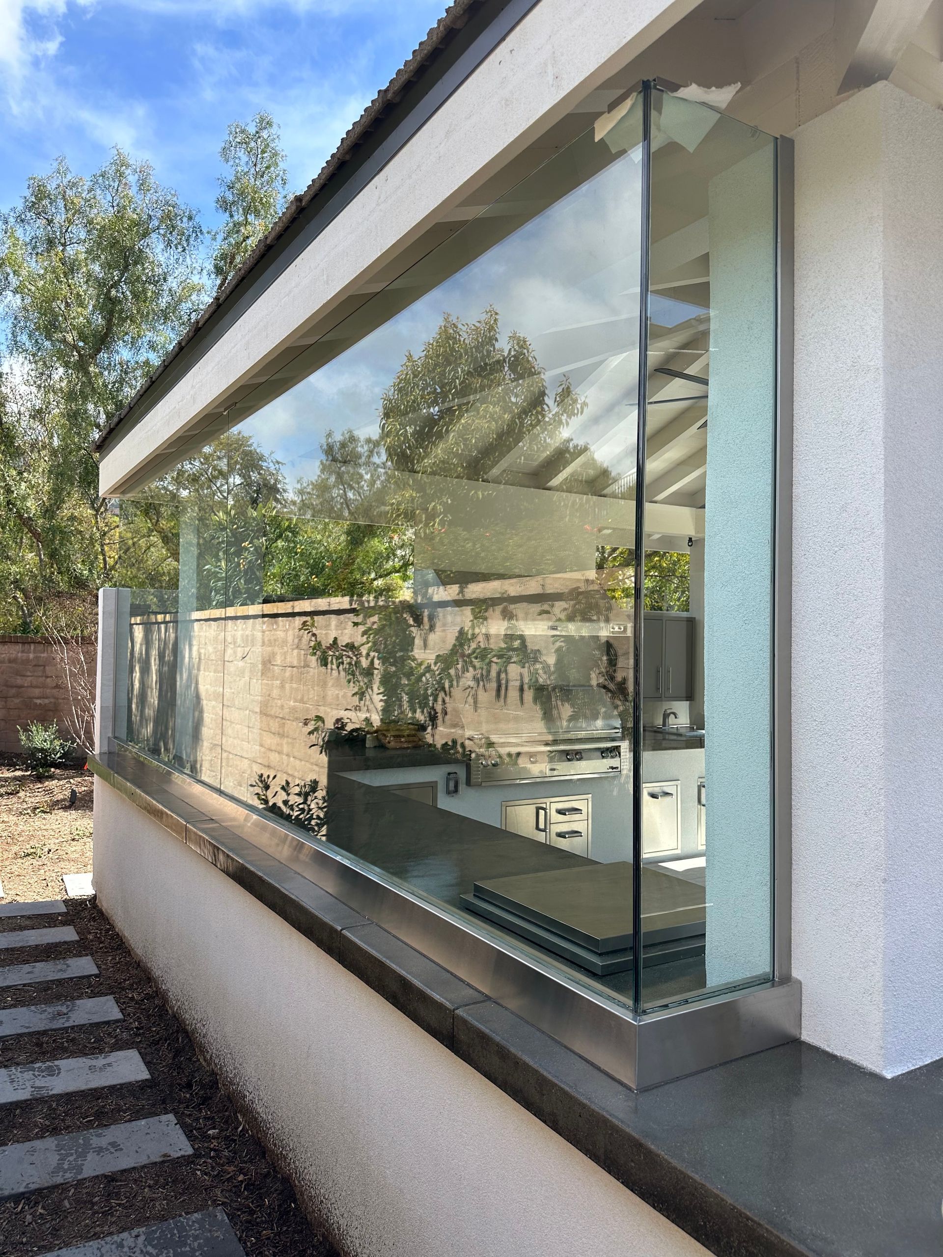 Glass wall of a modern building, showing a kitchen interior. Light stone and a paved pathway.