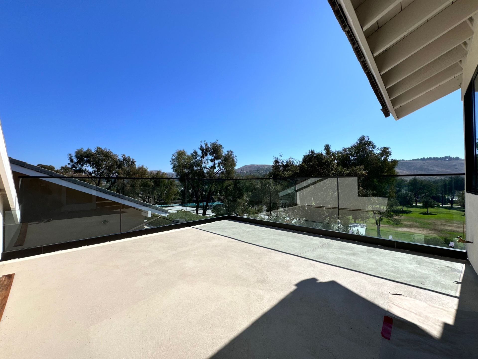 Empty rooftop patio with glass railings, overlooking a golf course and trees under a clear blue sky.