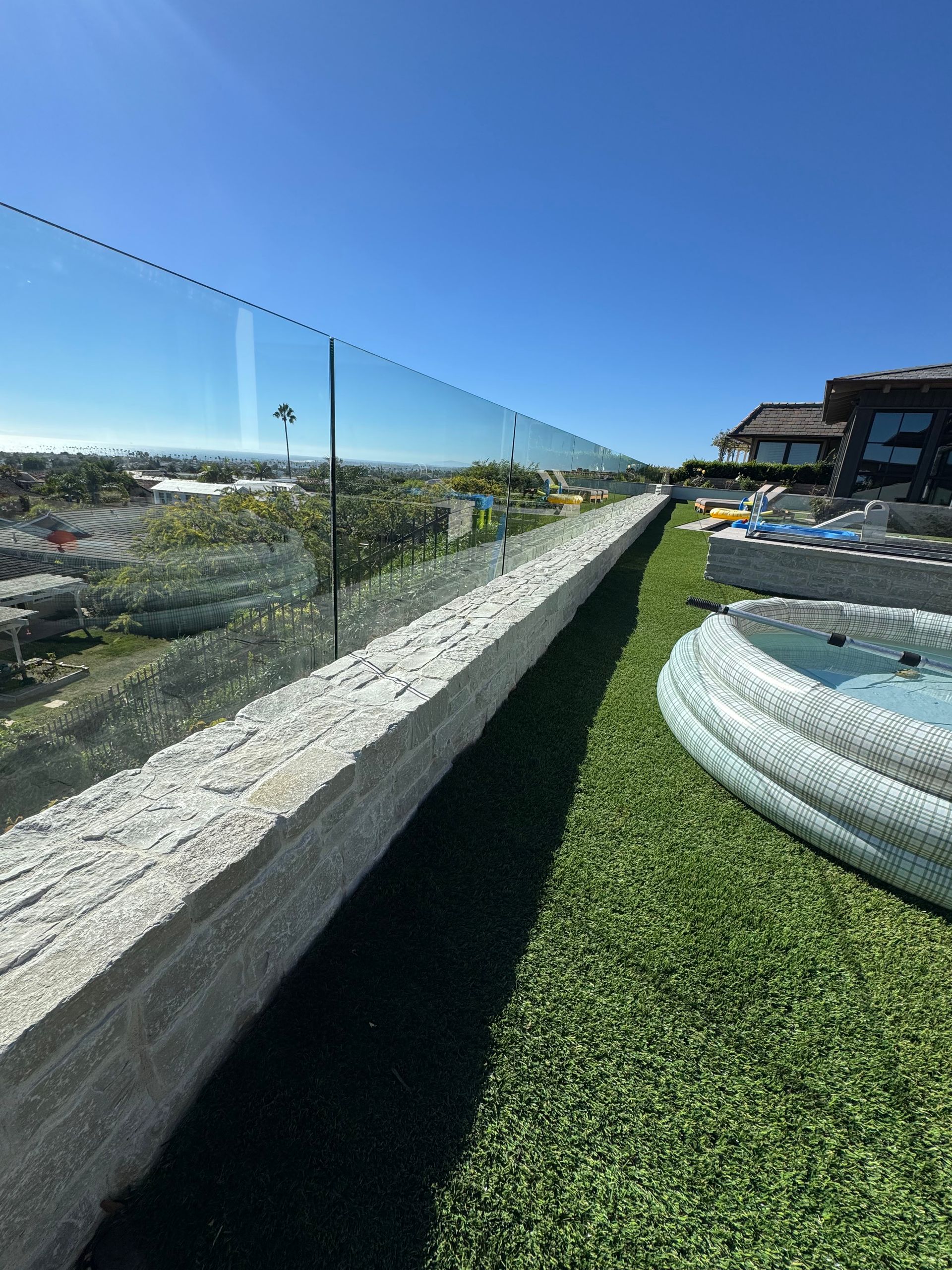 Long stone wall with glass railing, green turf, blue sky, and a view of distant buildings.