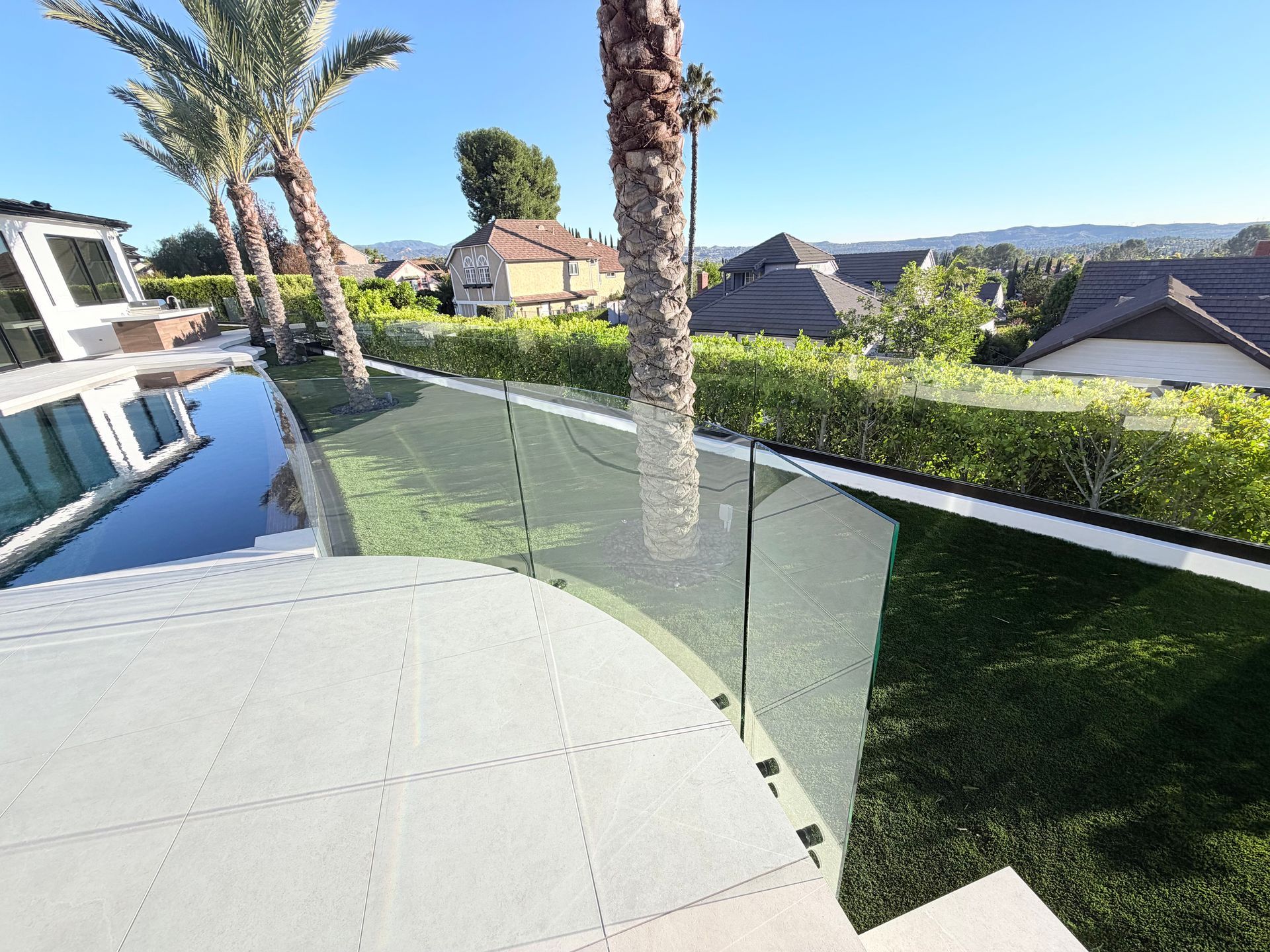 Poolside view with glass fence, palm trees, and houses in the background. Sunny day.