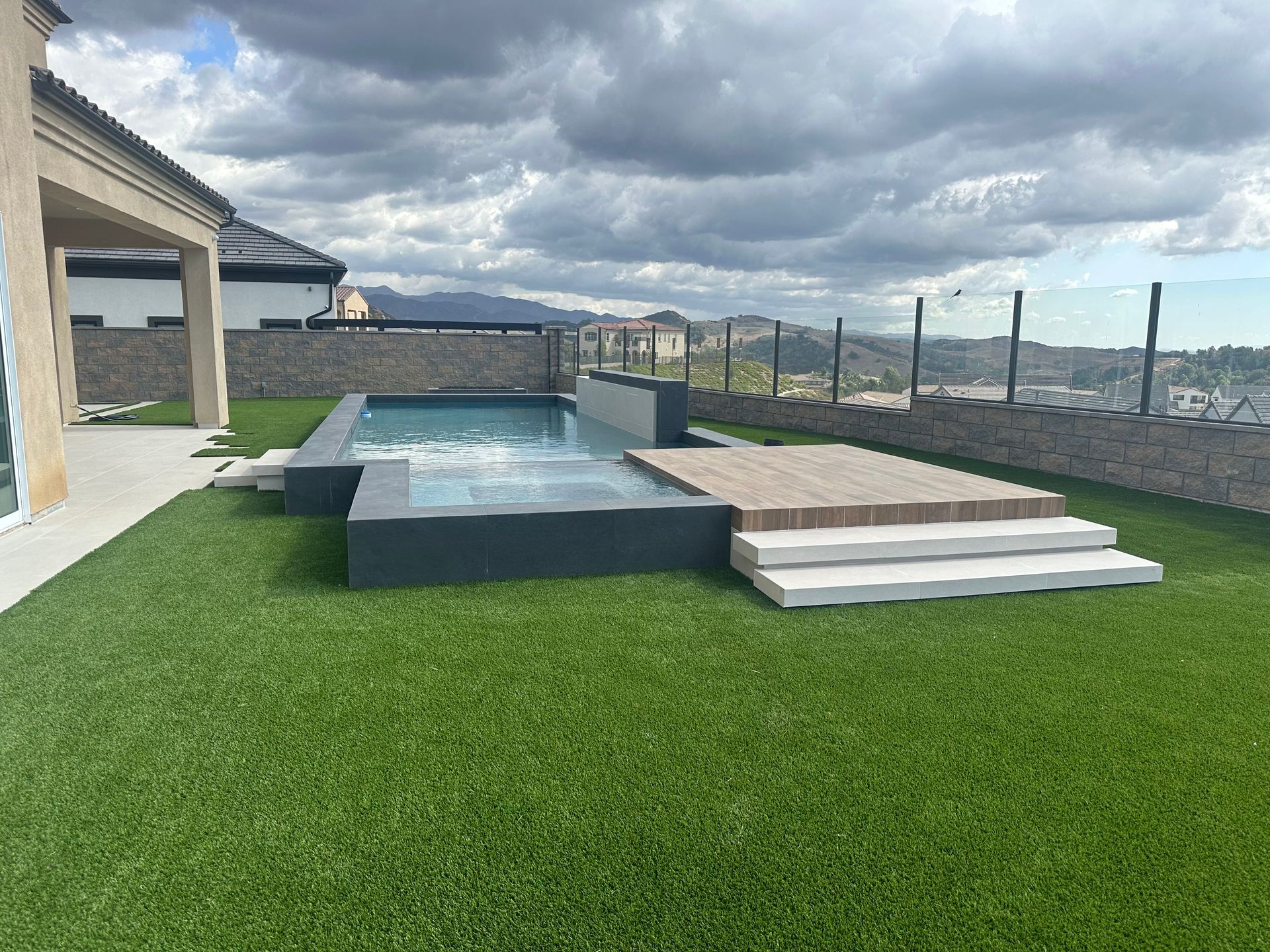 Backyard with rectangular pool, artificial turf, and a deck. Overcast sky, stone wall, and partial view of a building.