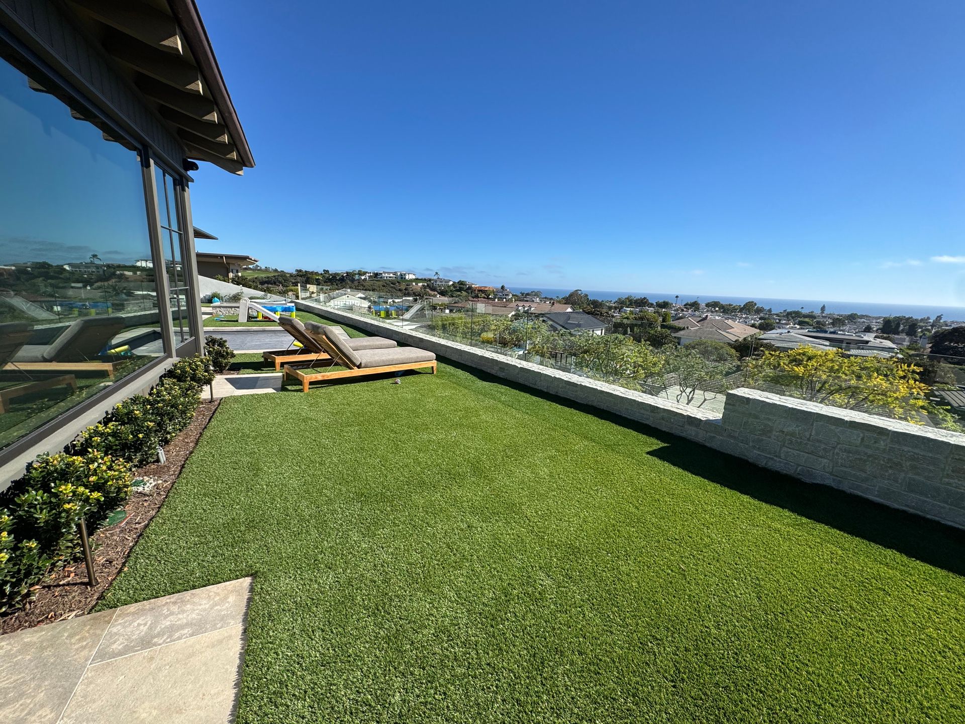 A rooftop patio with artificial turf, lounge chairs, and a panoramic ocean view under a clear blue sky.