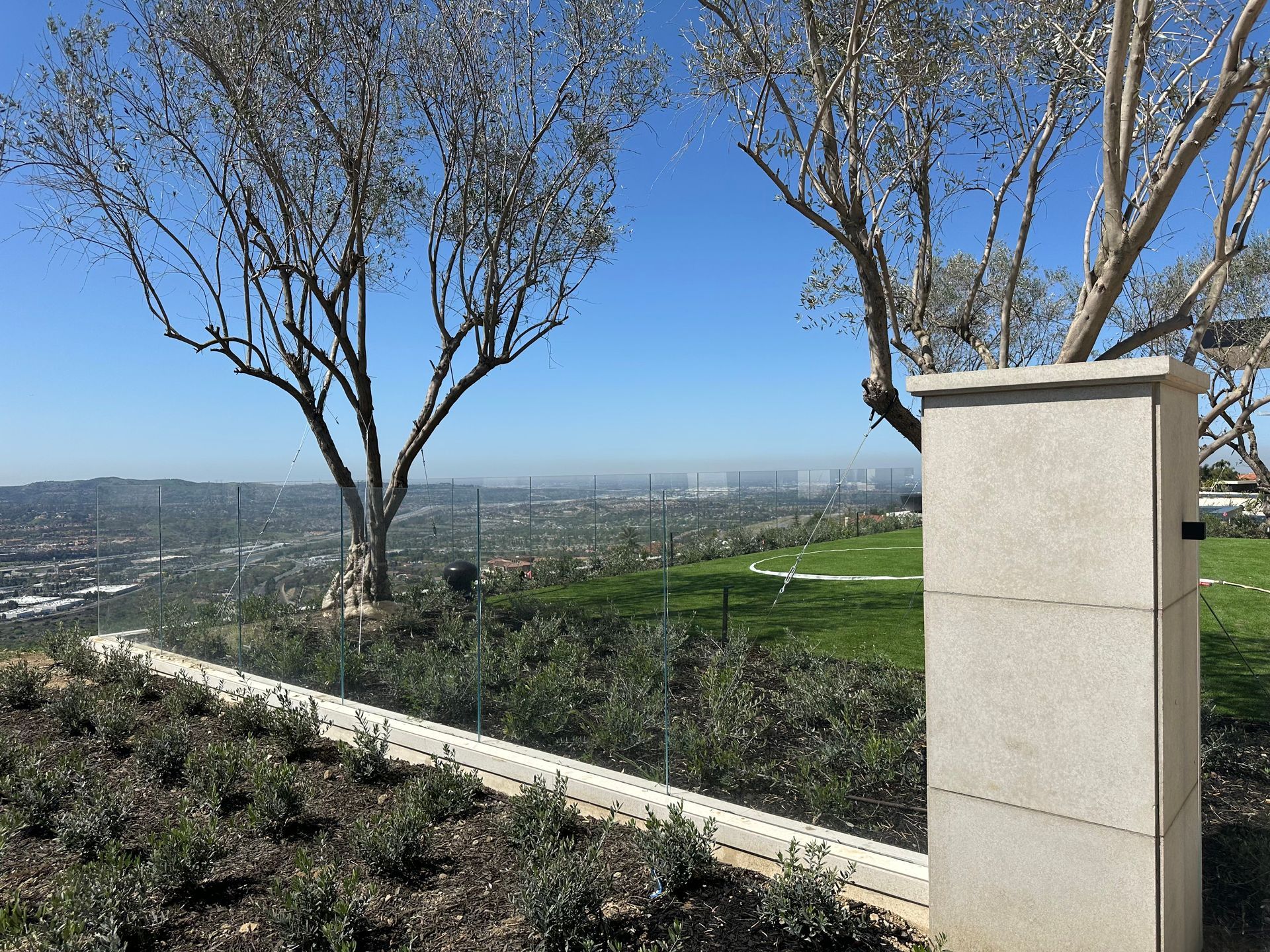 Scenic view overlooking a city from a hilltop, framed by trees and a stone pillar on a sunny day.