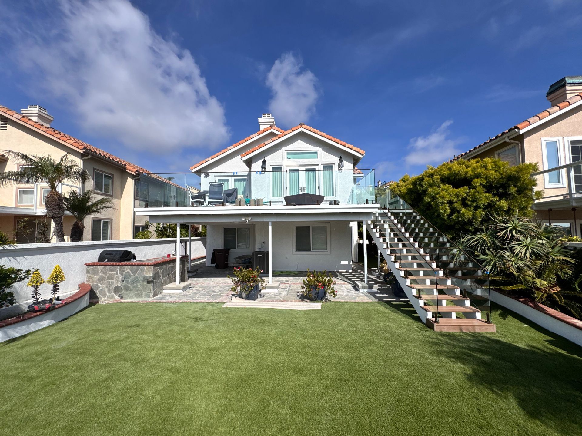 Backyard view of a two-story beach house with a rooftop deck, stairs, and a green lawn.