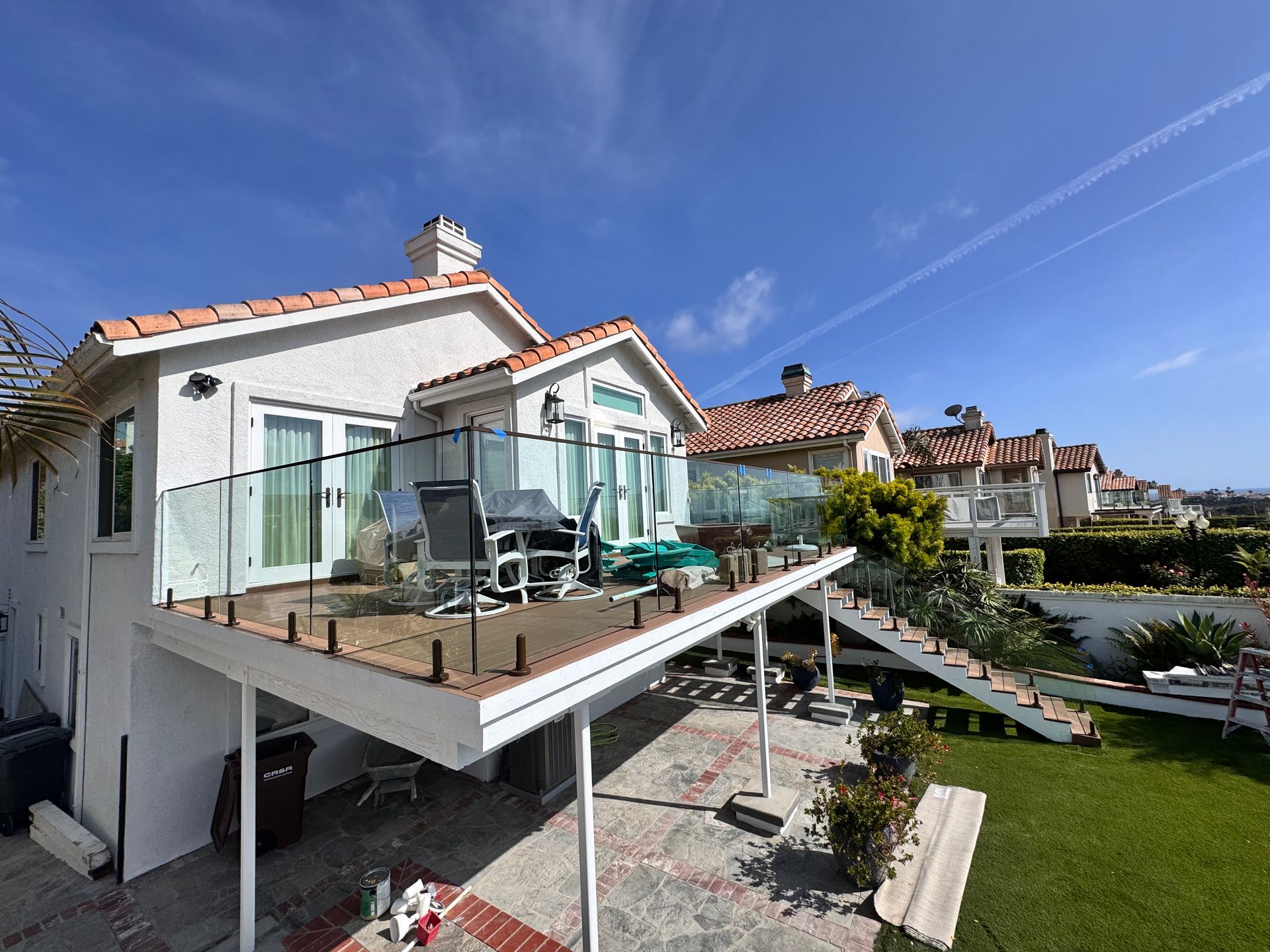 White stucco house with a wooden deck, glass railing, and tiled roof under a blue sky.