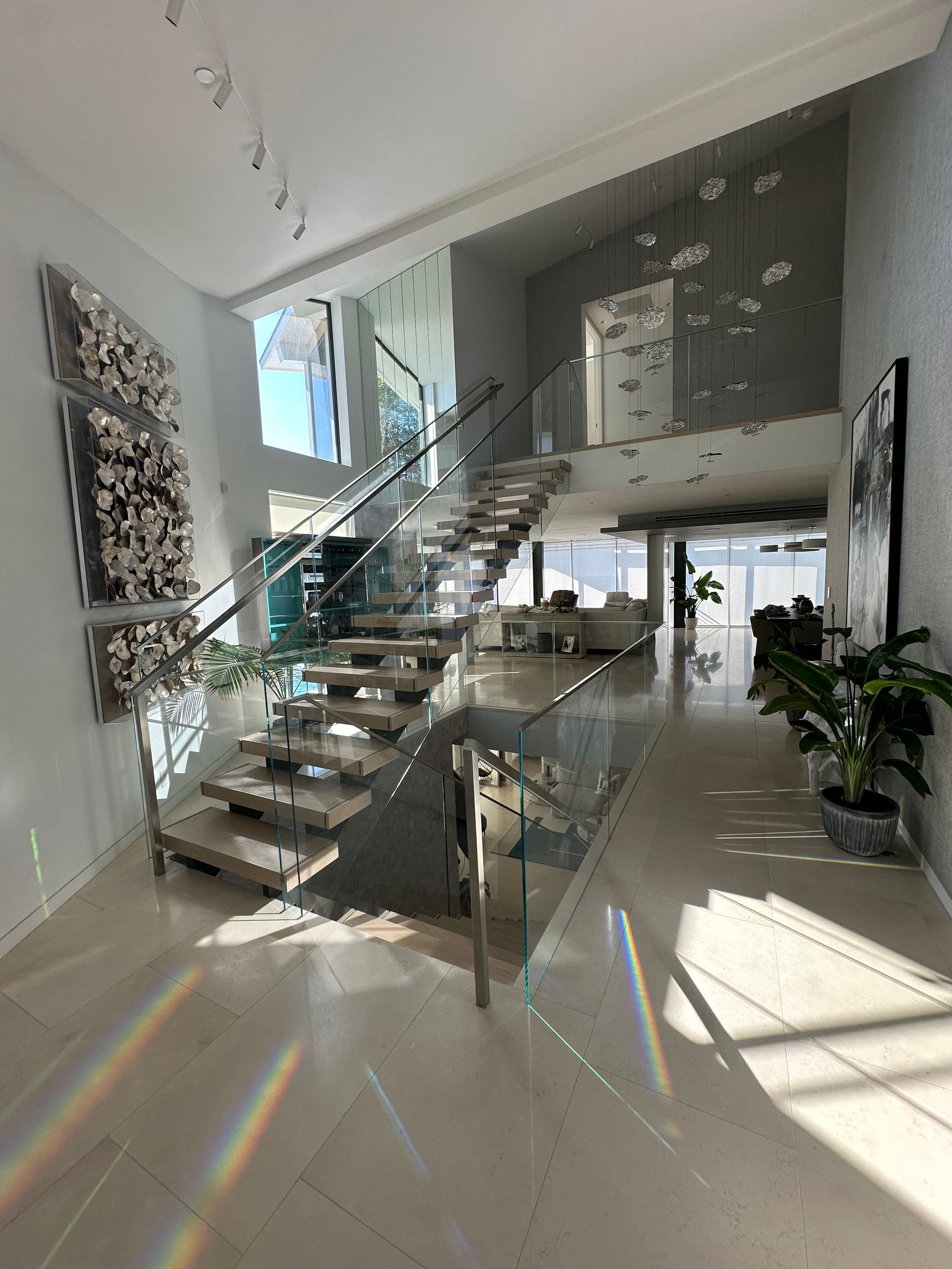 Modern, bright foyer with glass-railed staircase. Light streams across the tiled floor. Art pieces on walls.