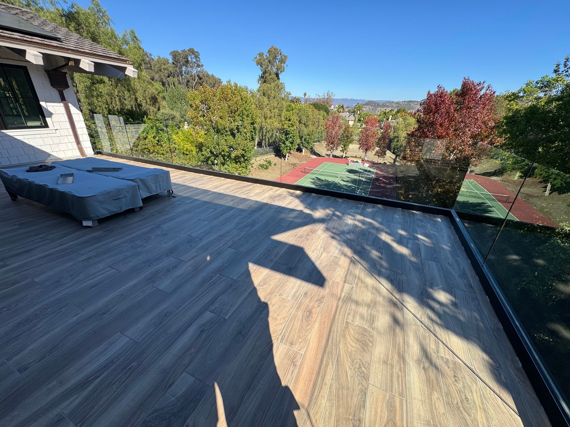 Wooden deck overlooking a tennis court and trees under a clear blue sky.