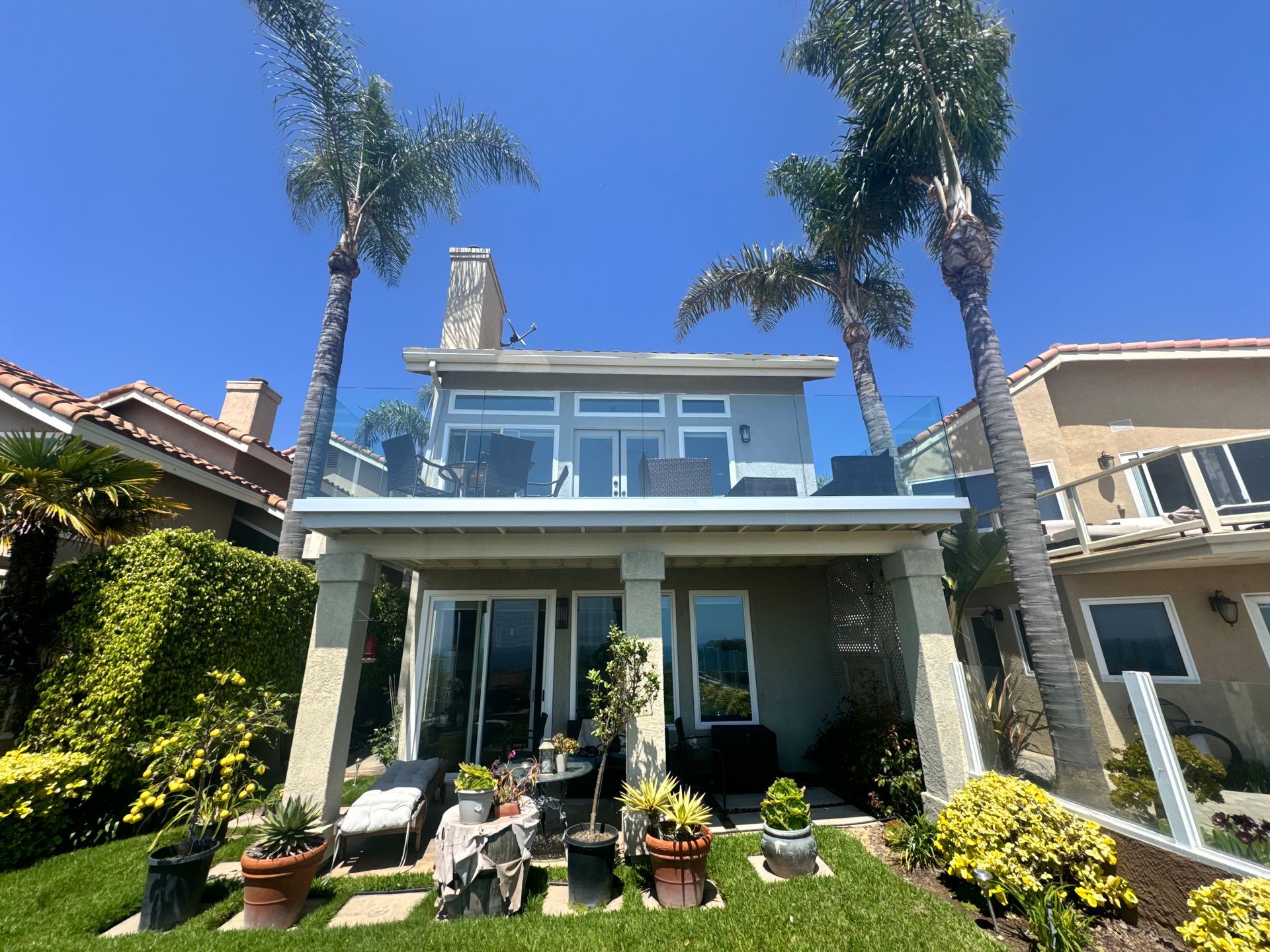 Two-story house with balcony and glass railings, palm trees, and green lawn under a blue sky.