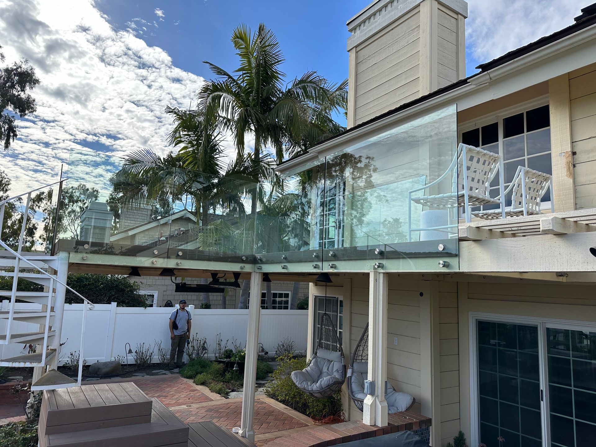A house with a glass balcony railing, with a person standing below on a patio with greenery.