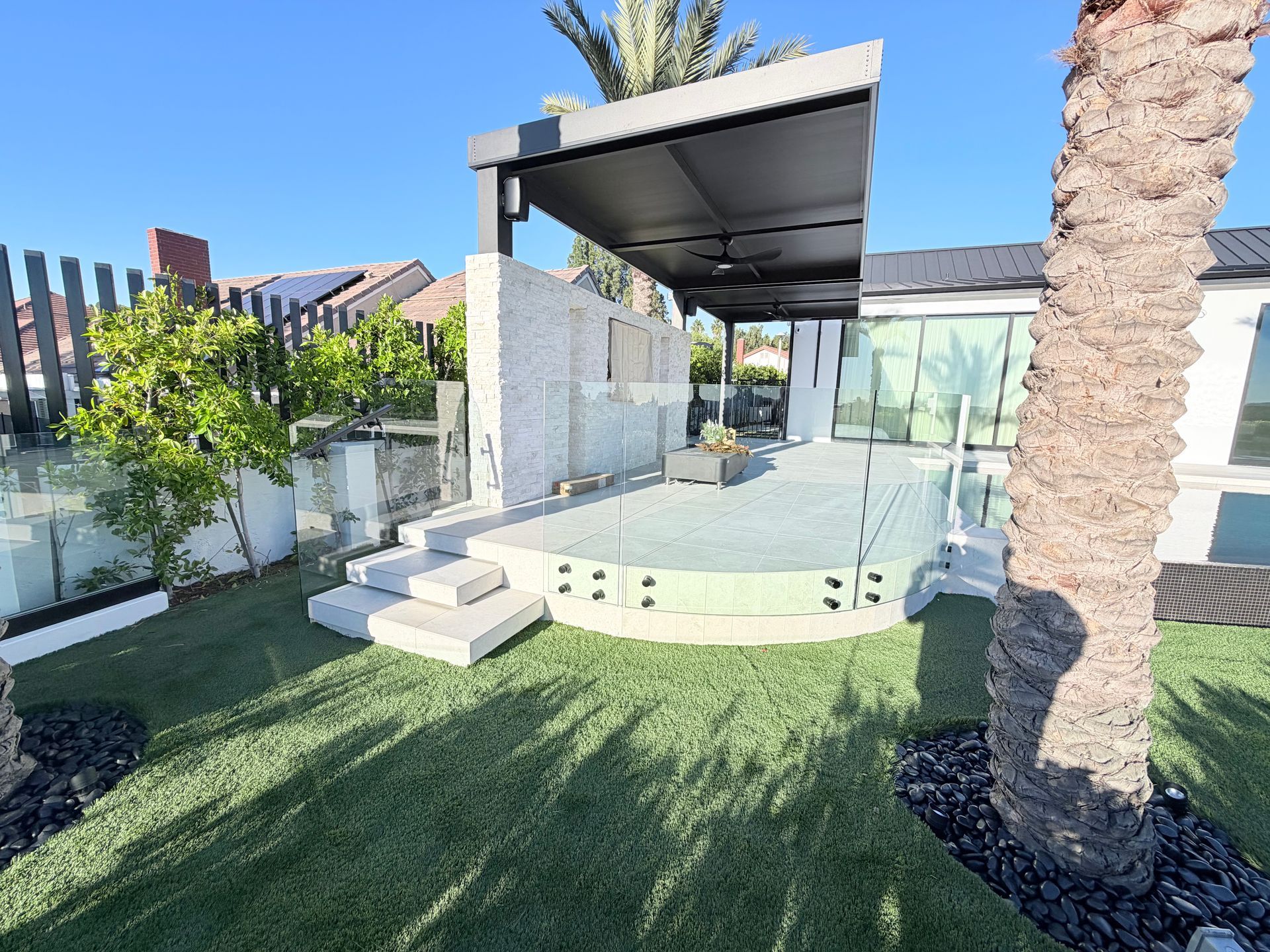 Backyard patio with modern design, stone wall, pergola, steps, artificial grass, and palm trees.