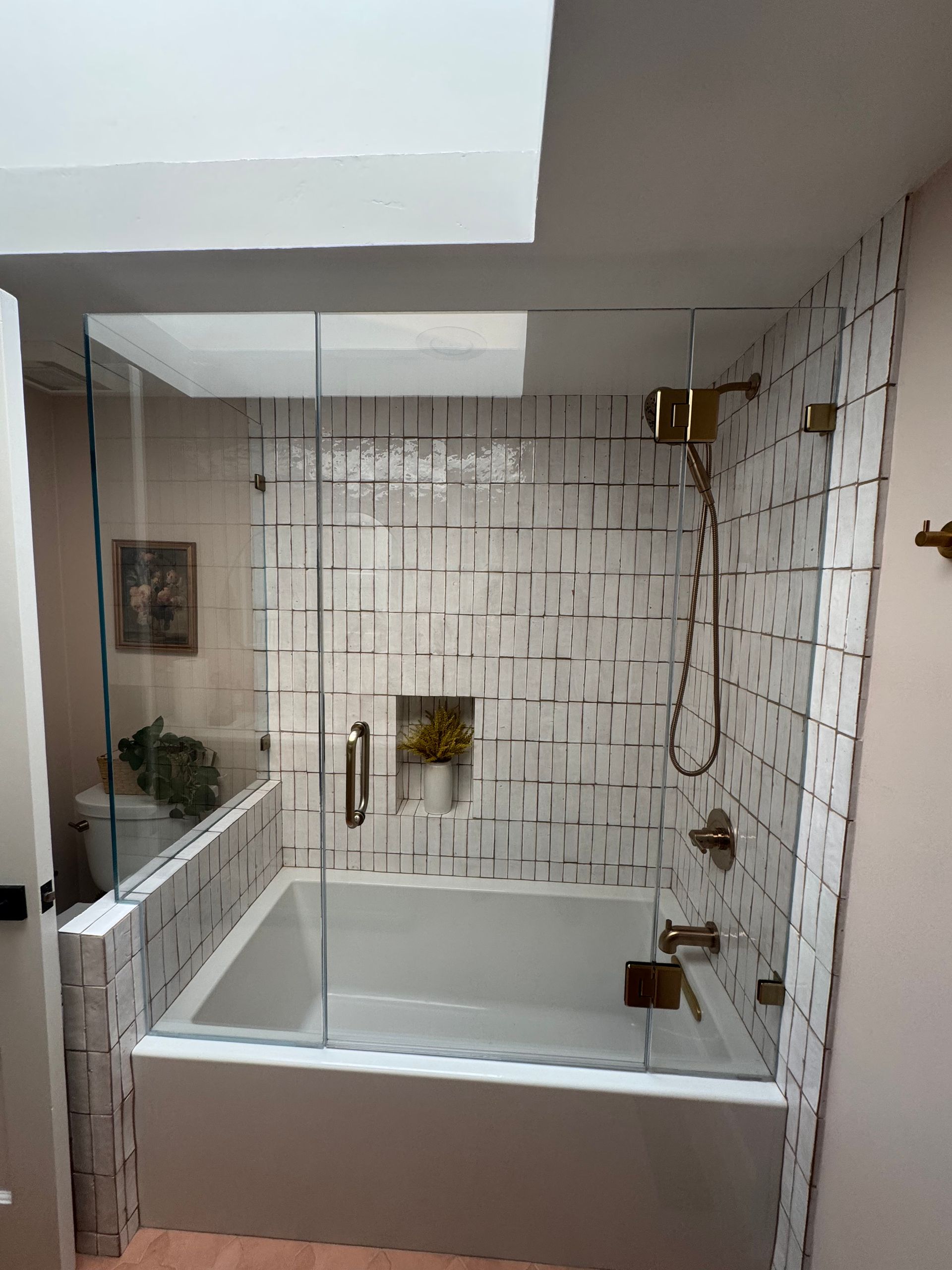 Bathroom with white tub, gold fixtures, and patterned tile. Glass shower door and skylight are visible.