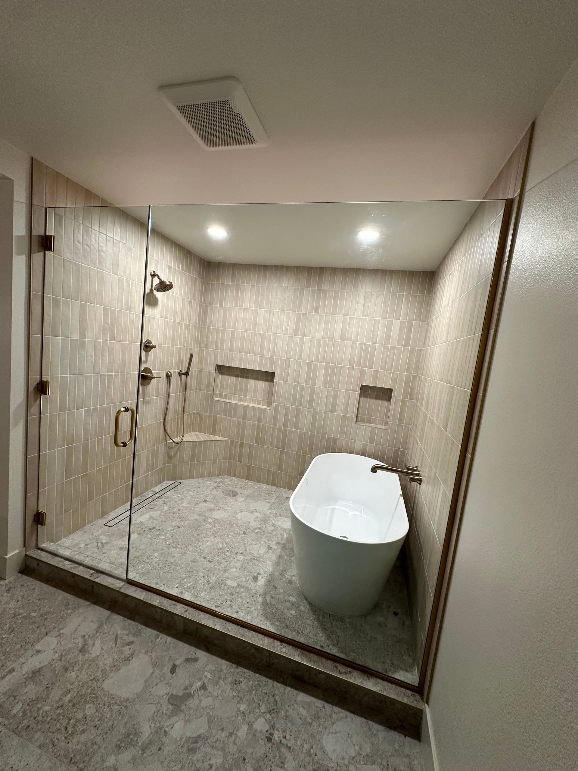 Modern bathroom with glass shower, tub, beige tile, and pebble floor.