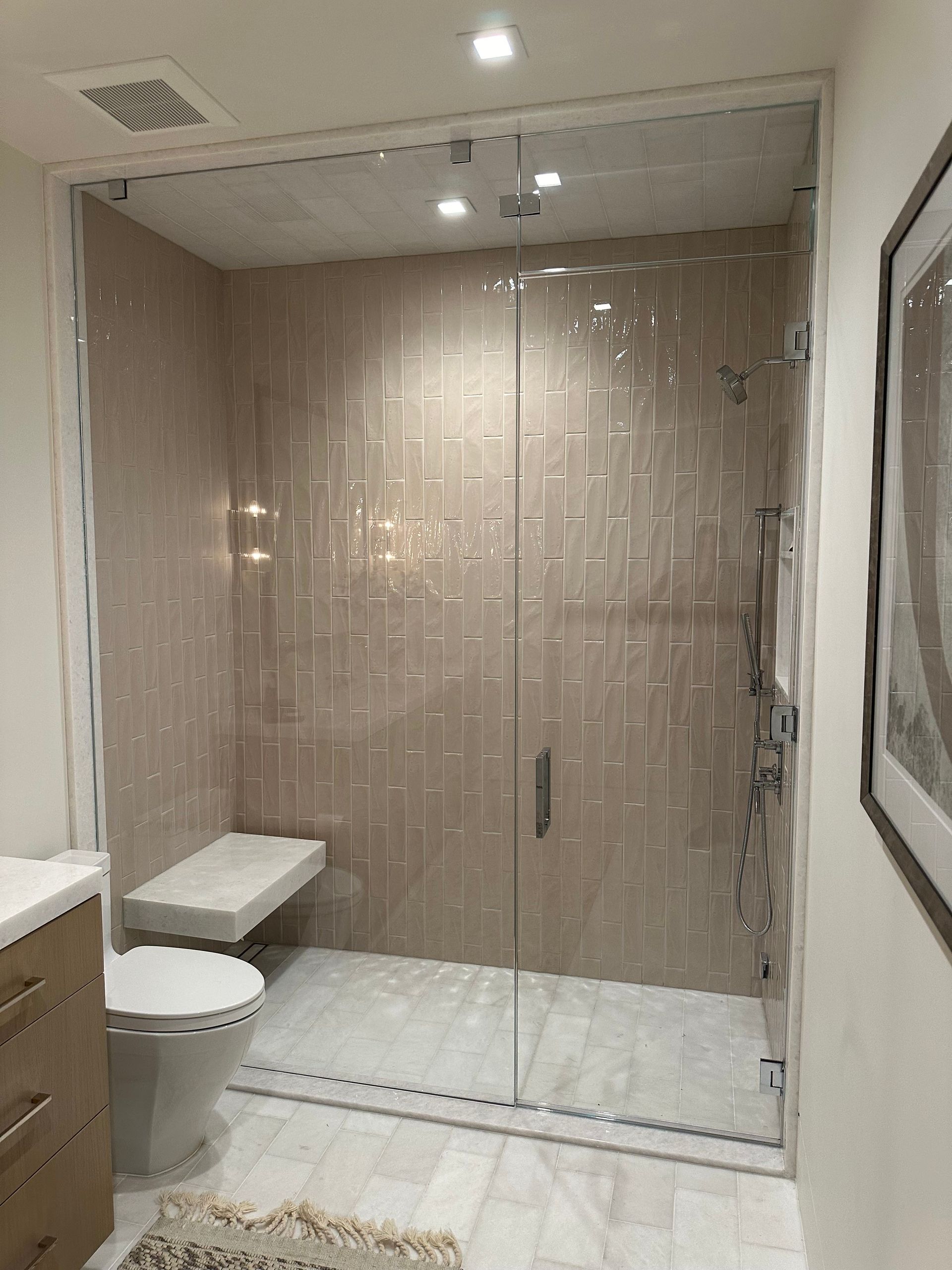 Modern bathroom with a glass-enclosed shower, beige tile, toilet, and a small bench.