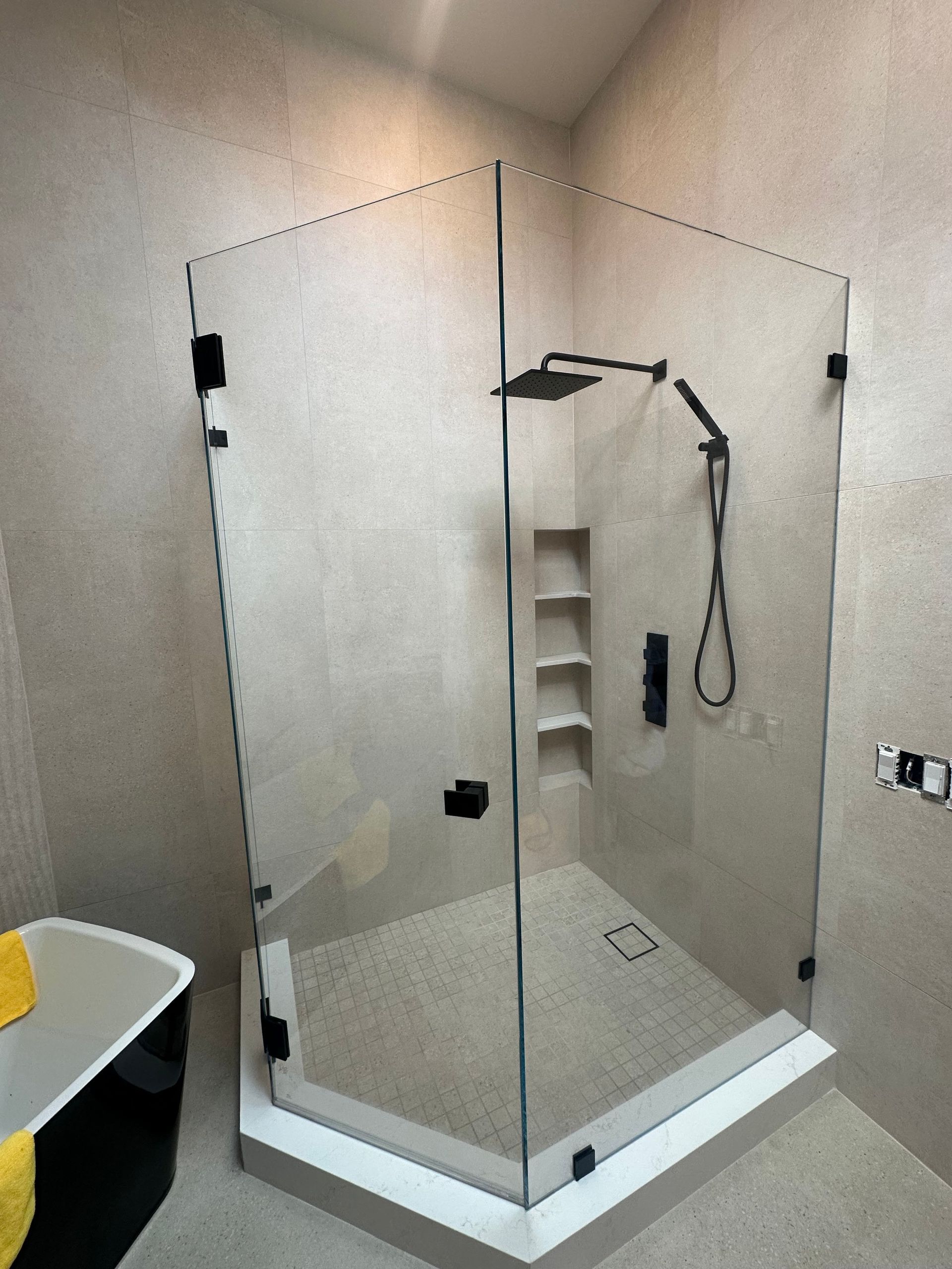 Modern glass corner shower with black fixtures and recessed shelving, on a light tile base.