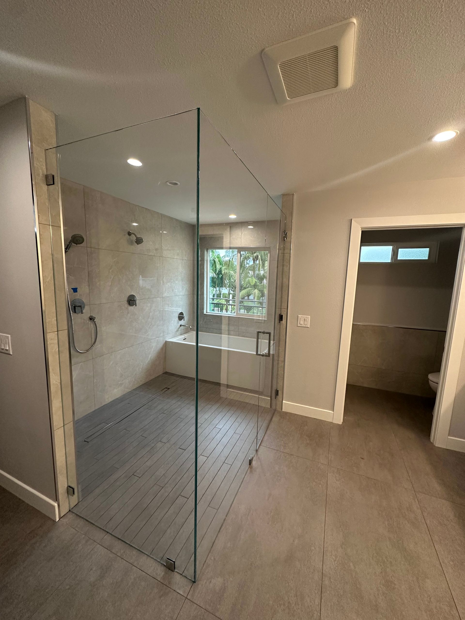 Modern bathroom with glass shower enclosure, tiled walls, and gray flooring. Bathtub visible through a window.