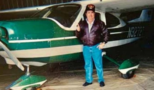 A person wearing a jacket and hat stands next to a green and white light aircraft inside a hangar.