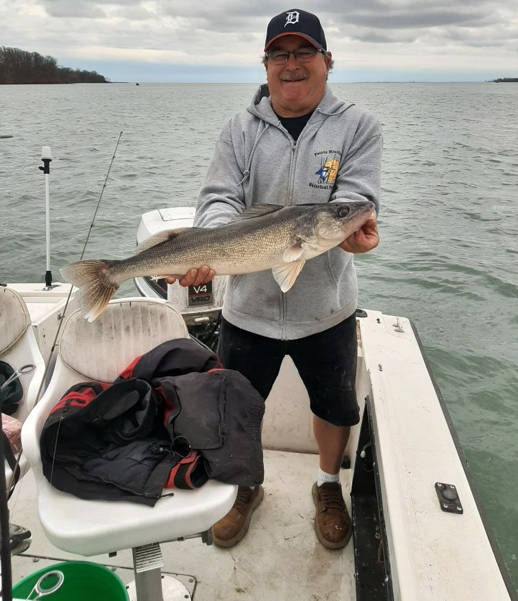 A smiling person in a Detroit Tigers hat and grey hoodie holds a large fish while standing on a boat on a lake.