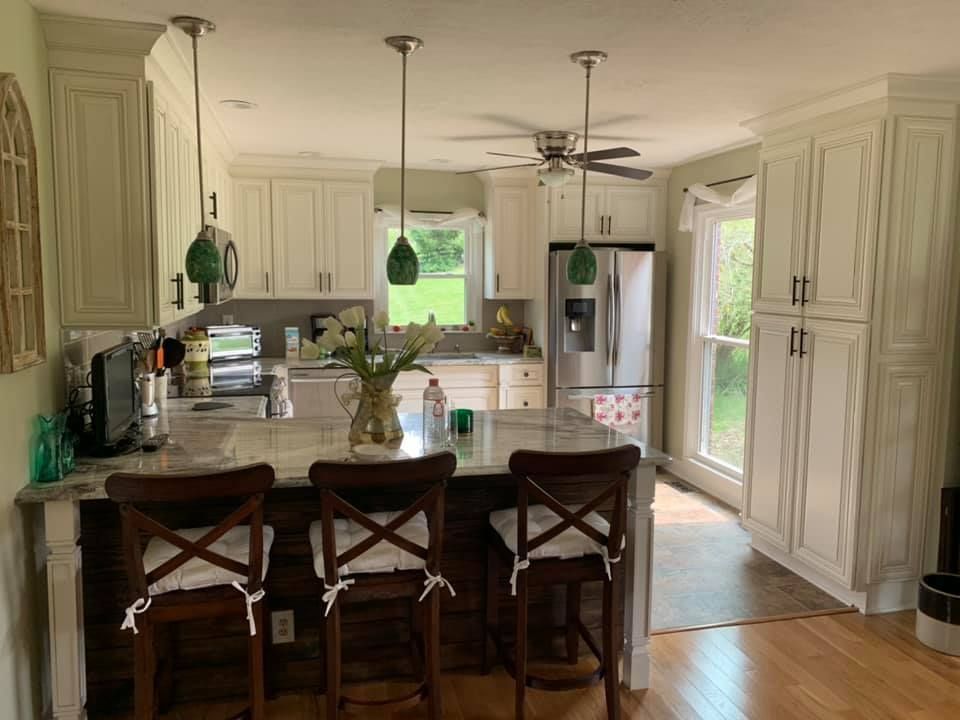 a kitchen with white cabinets , stools , a refrigerator and a ceiling fan .